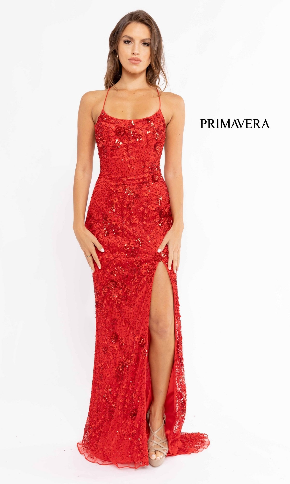 A woman modeling the Strappy-Back Sequin Long Prom Dress 3931, featuring red sequins, spaghetti straps, and a thigh-high slit, stands against a white background with the “PRIMAVERA” logo beside her.