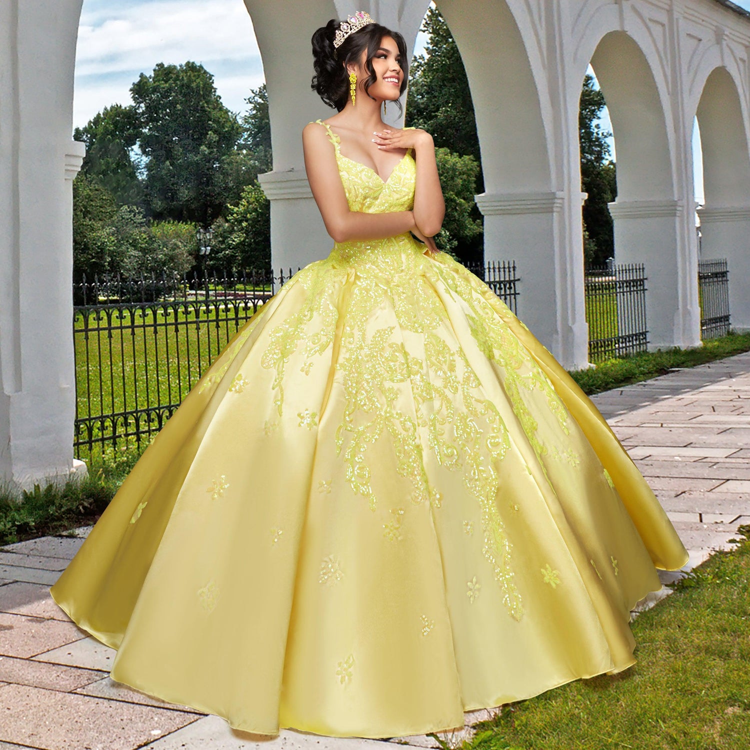 A woman smiles with her hands near her face, standing outdoors before white arches, wearing the yellow, floral-embroidered Quinceañera Sample Dress QF106 with a v-neckline and a tiara.