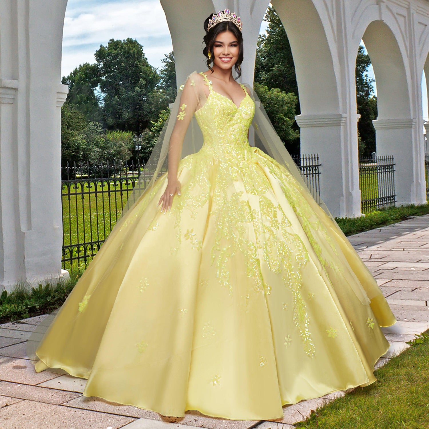 A woman stands outdoors near white arches and a black iron fence, wearing the Quinceañera Sample Dress QF106—a yellow v-neck gown—along with a tiara and veil, surrounded by trees and grass.
