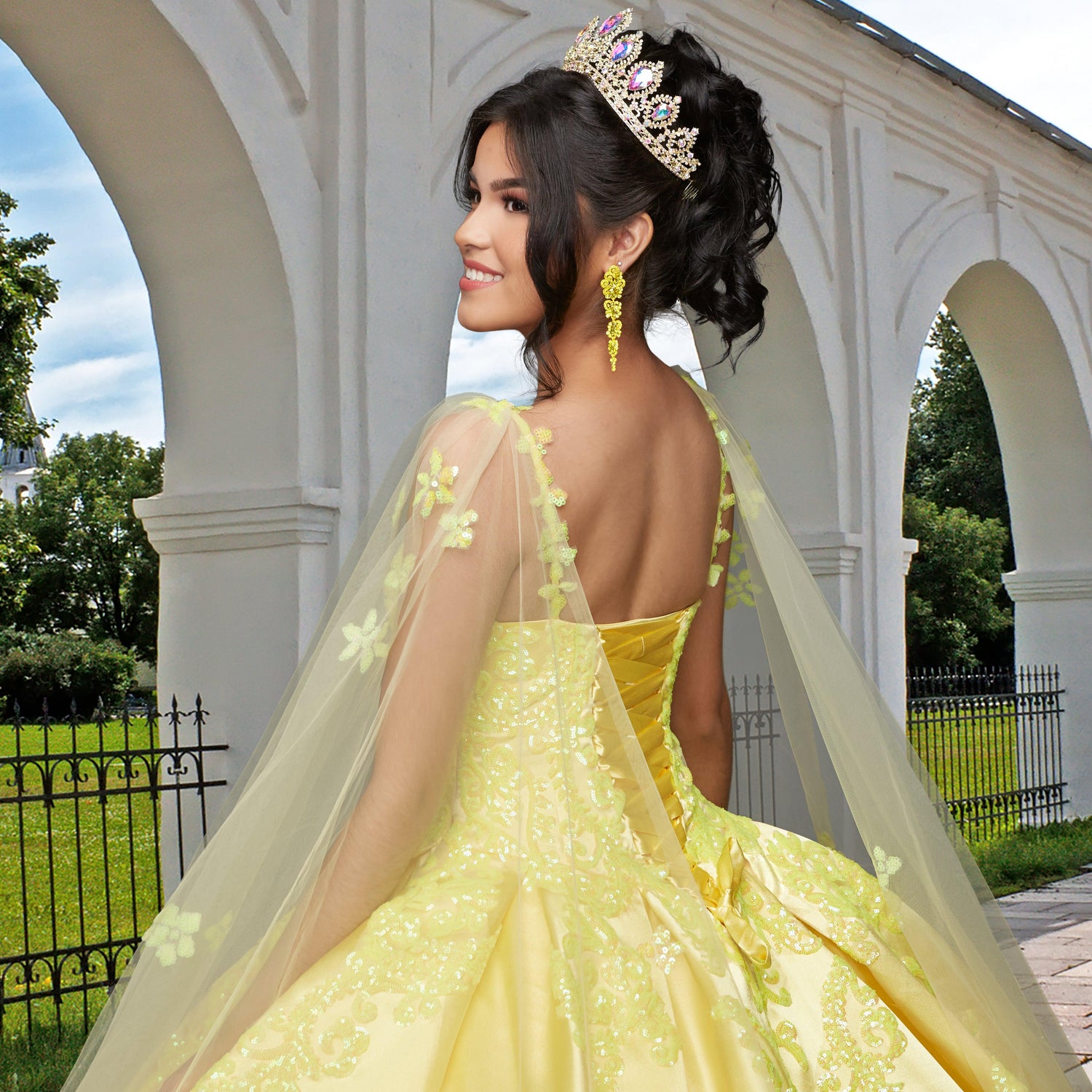 A young woman wears the Quinceañera Sample Dress QF106, a yellow v-neck gown, and a tiara, smiling outdoors near white arches and greenery.