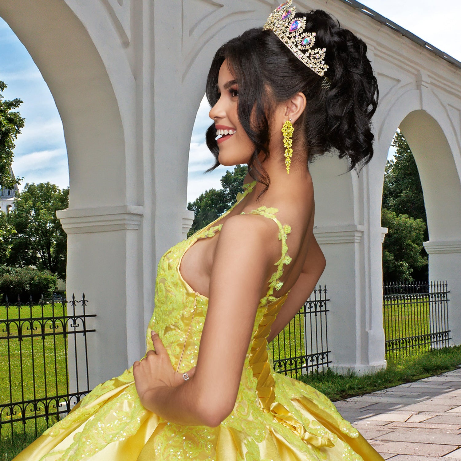 A young woman wears the Quinceañera Sample Dress QF106, a yellow sequin gown with a tiara, smiling outdoors with her hands on her hips in front of a white arch and lush greenery.