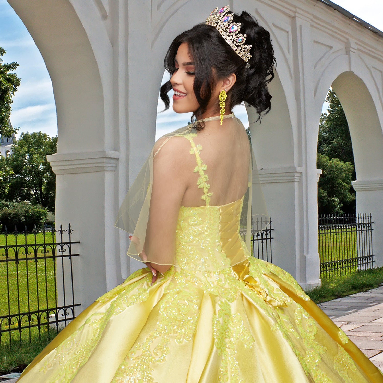 A young woman wearing the Quinceañera Sample Dress QF106 and a tiara stands outdoors by an arched white structure, smiling over her shoulder.