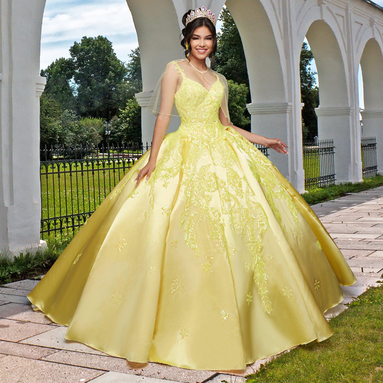 A young woman in the Quinceañera Sample Dress QF106 and a tiara stands outdoors on a stone path beside white arches and a black fence.