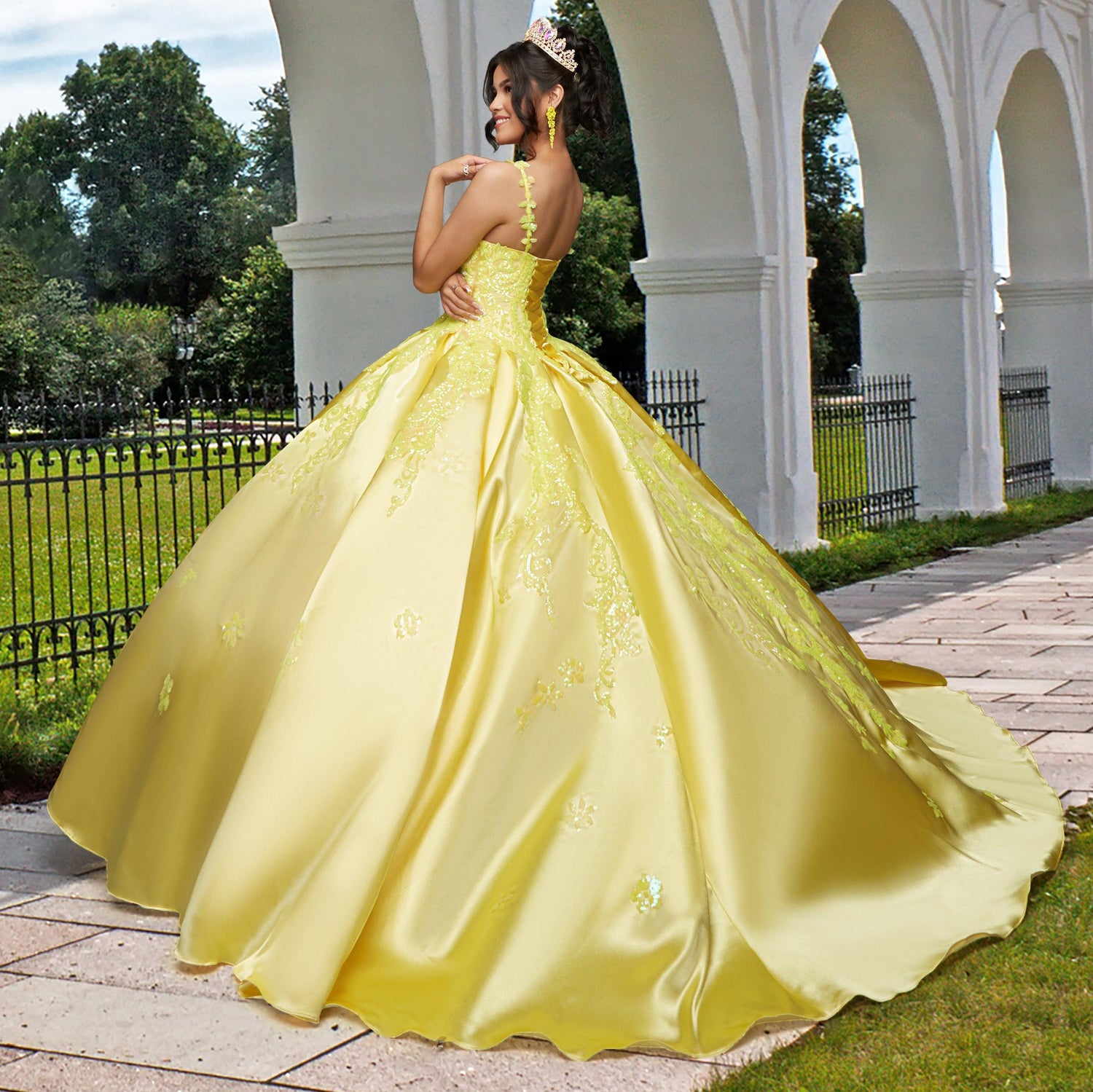 A woman wearing the Quinceañera Sample Dress QF106 and a tiara stands outdoors on a stone path by white arches and a black iron fence.