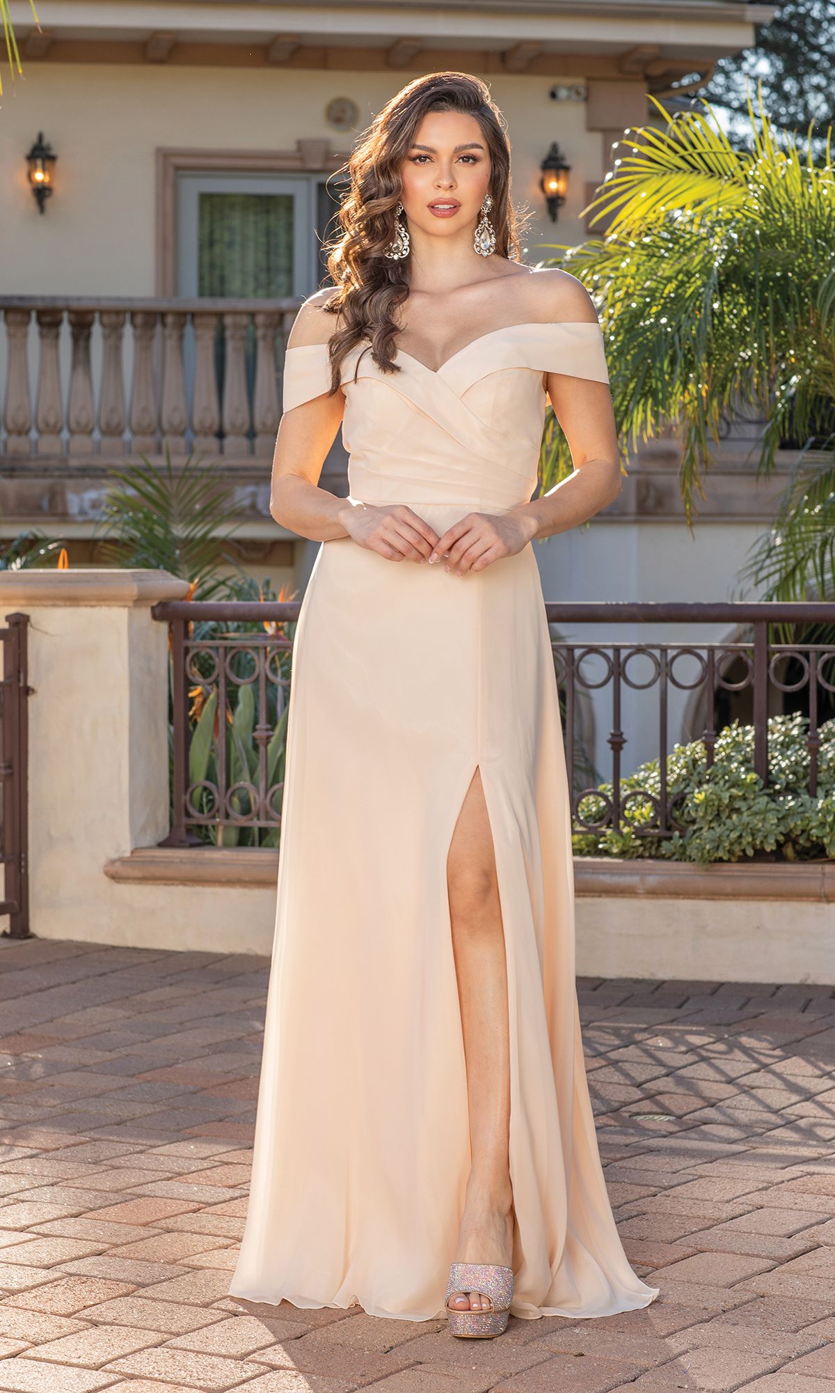 A woman in the Off-the-Shoulder Classic Formal Prom Dress with Slit, in light peach, poses on a paved patio before a building adorned with a balcony, lush plants, and elegant iron railing.
