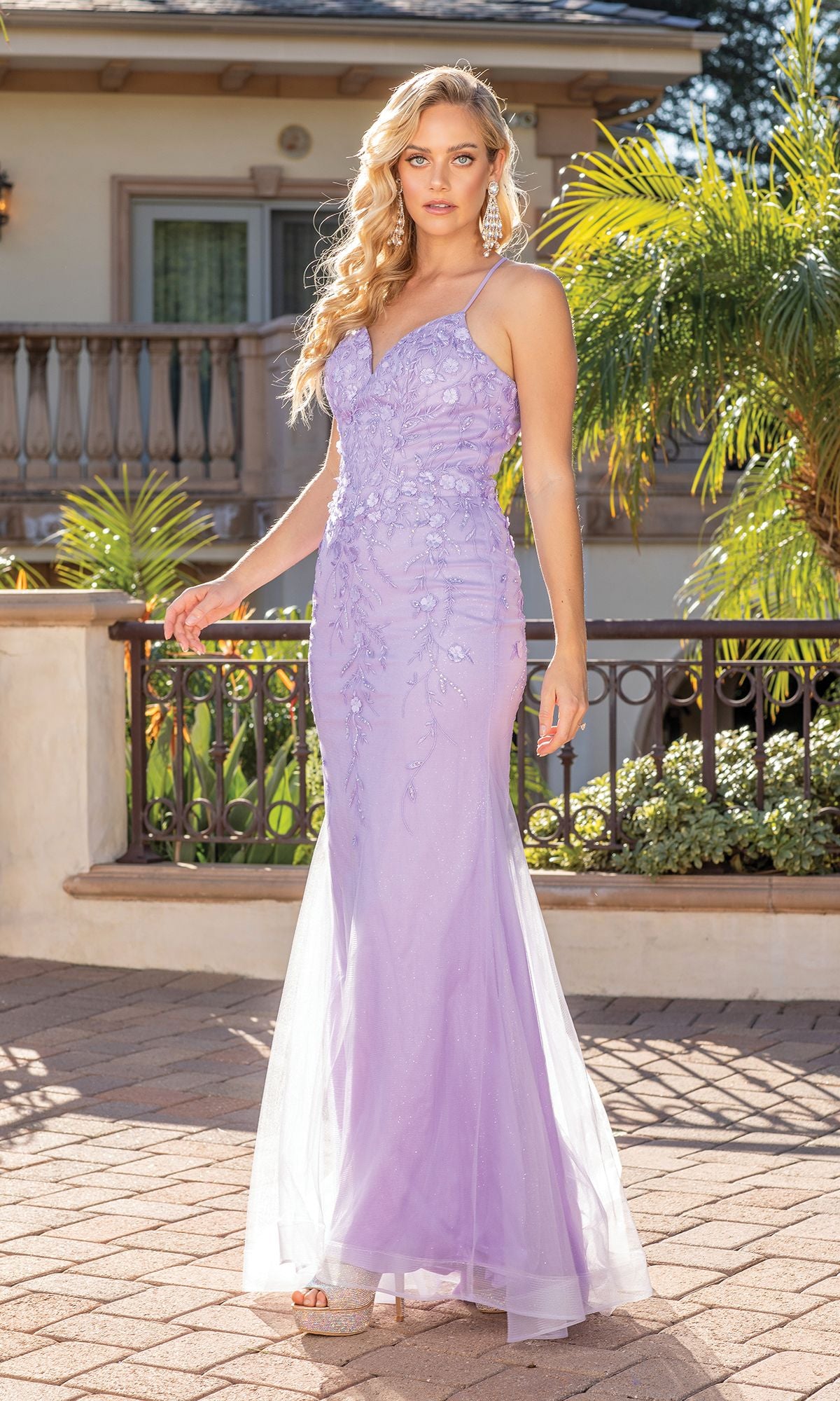 A woman stands outdoors by a balcony and palm trees, wearing the Glitter V-Neck Prom Dress with 3-D Floral Appliques in light purple.