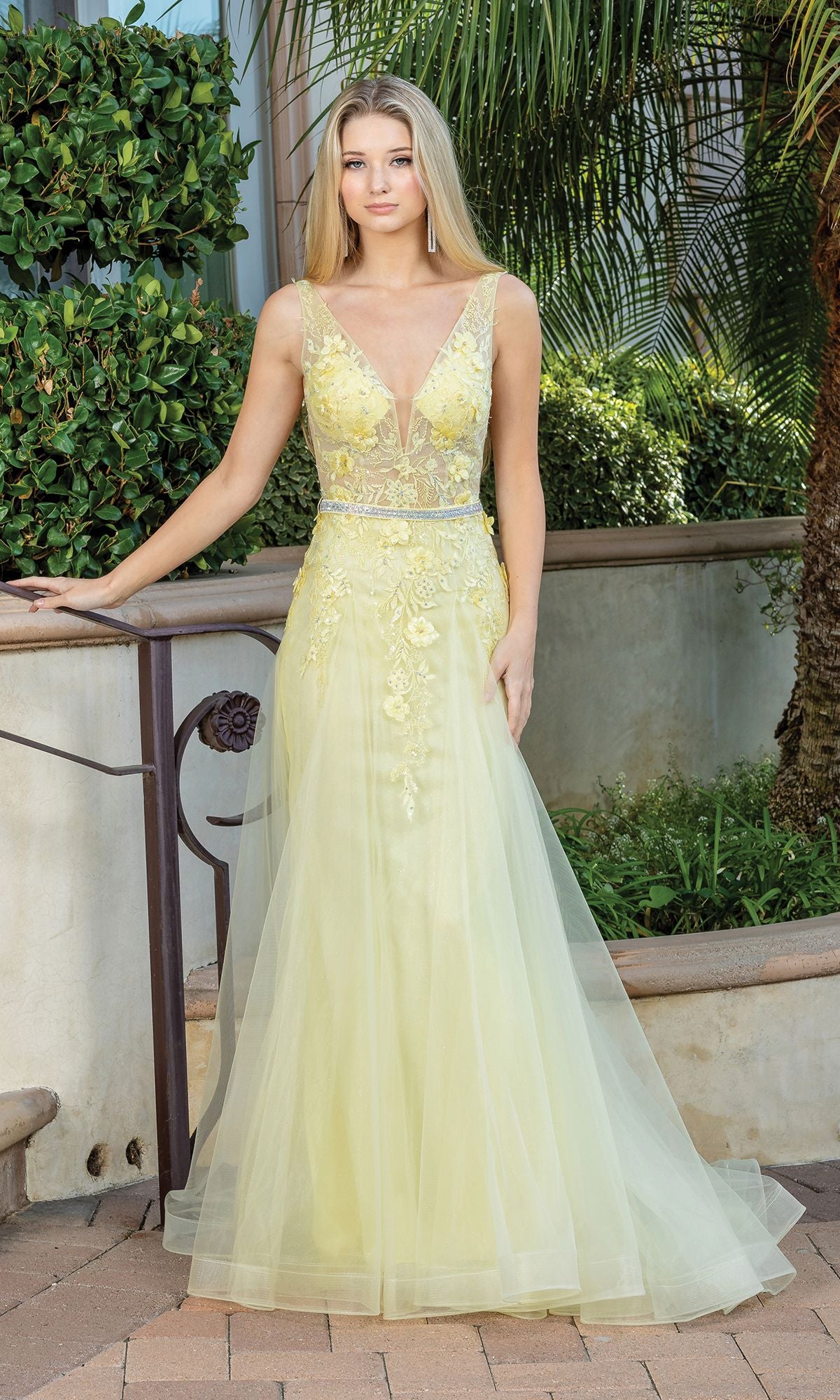 A woman stands outdoors wearing the V-Neck Prom Ball Gown with Sheer Bodice, a sleeveless, floor-length yellow dress featuring beaded floral appliques and a deep V neckline.