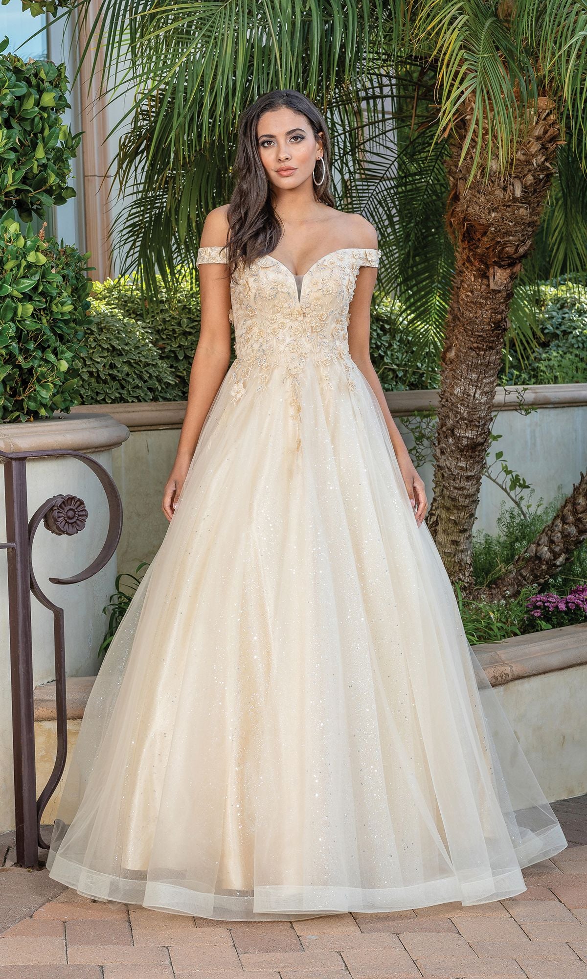 A woman wearing the Off-Shoulder Prom Ball Gown with Corset Back, a cream ball gown featuring floral embellishments, stands outdoors near lush greenery and palm trees.