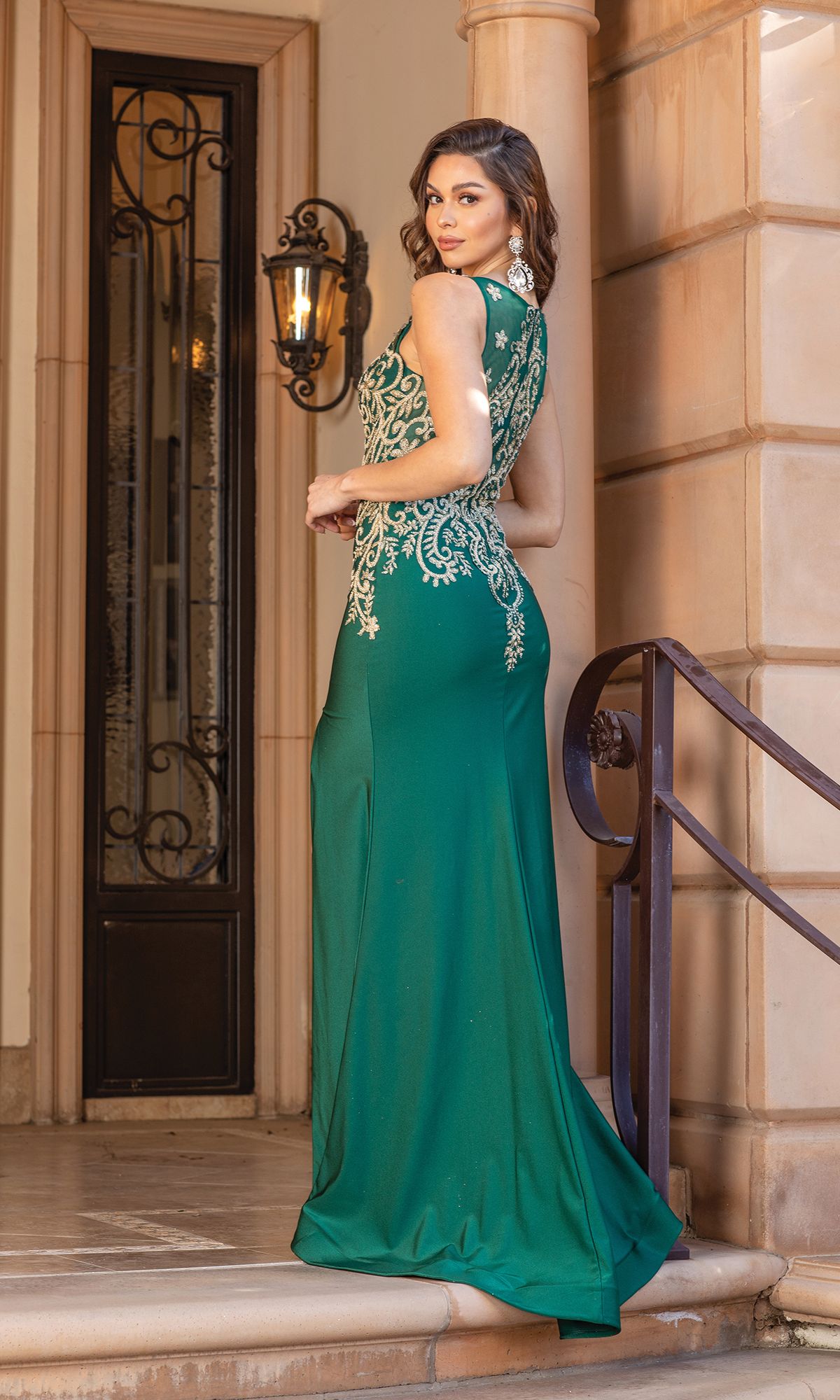 A woman wearing the Sleeveless V-Neck Gold-Embroidered Prom Dress 4325 stands on stairs, looking over her shoulder in front of a building with tan stone walls and a decorative door.