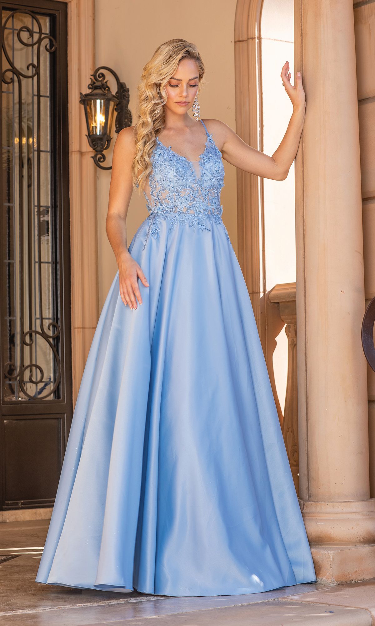 A woman in the A-Line Long Prom Ball Gown: Dancing Queen 4326, featuring a light blue hue and lace bodice, stands by a stone pillar outdoors, capturing the elegance of a Dancing Queen.