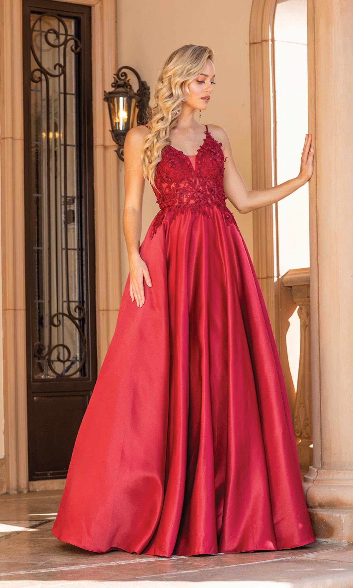 Wearing the Dancing Queen 4326 A-Line Long Prom Ball Gown, a woman stands by a column in an elegant setting, lace detailing enhancing her timeless grace as she looks to the side.