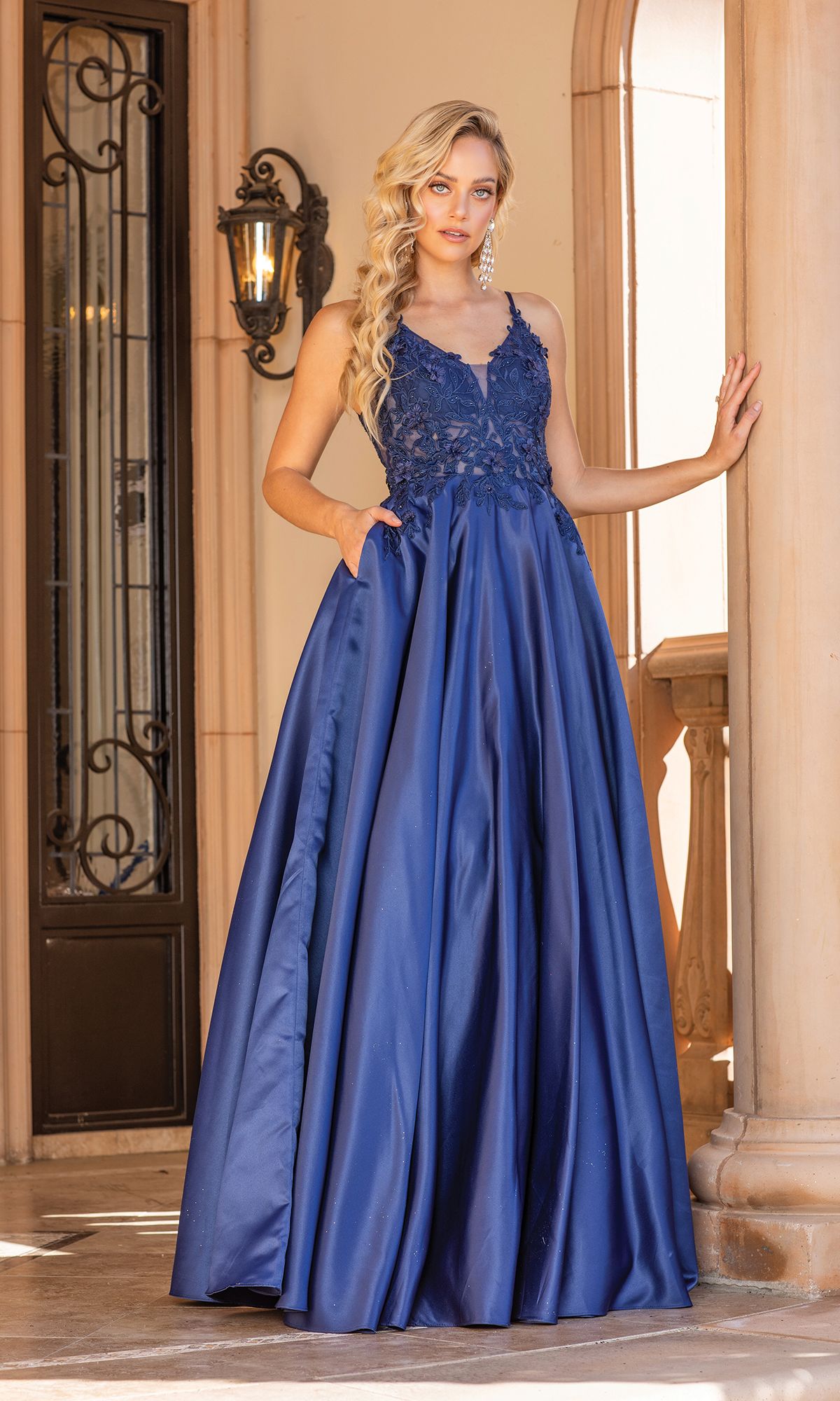 A woman in the Dancing Queen 4326 A-Line Long Prom Ball Gown, featuring blue satin and lace detailing, stands beneath a stone archway with one hand resting on a column.