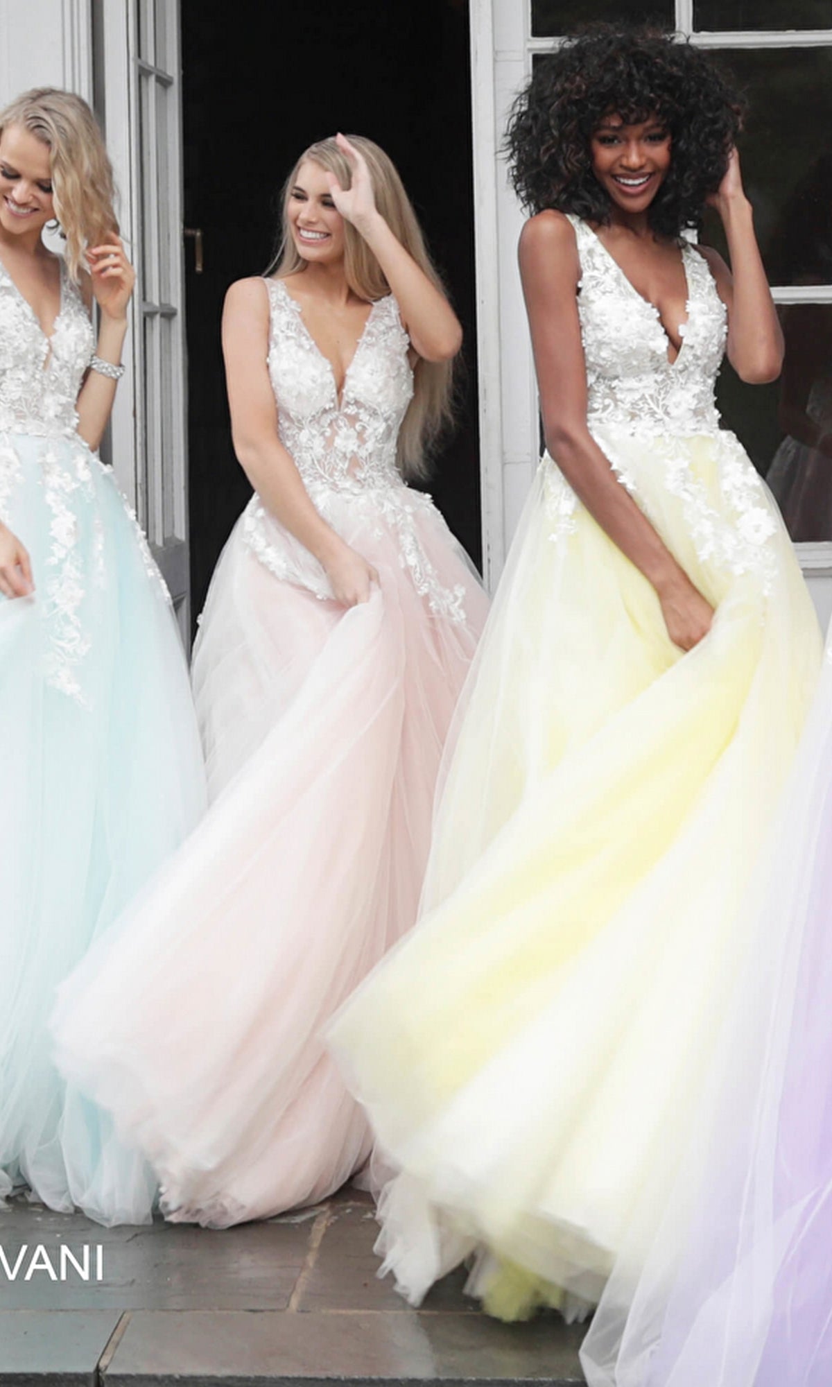 Three women stand smiling in pastel Jovani Tulle Prom Ball Gowns with floral appliques and lace bodices, posing together in front of a doorway.