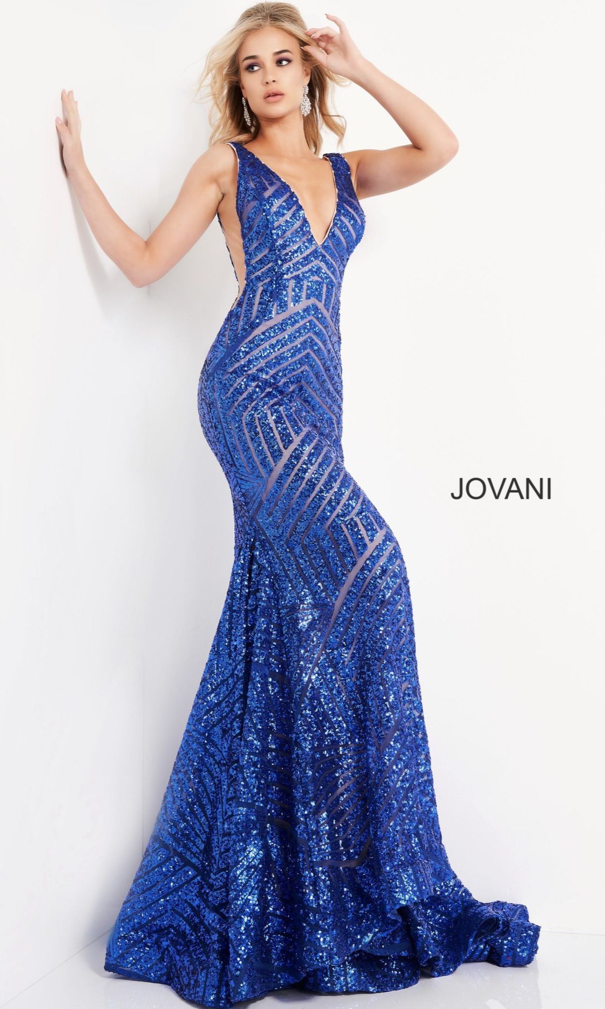 A woman poses against a white wall in the Jovani Plus-Size Mermaid Prom Dress with Sequins, a sleeveless, floor-length gown featuring blue geometric patterns.