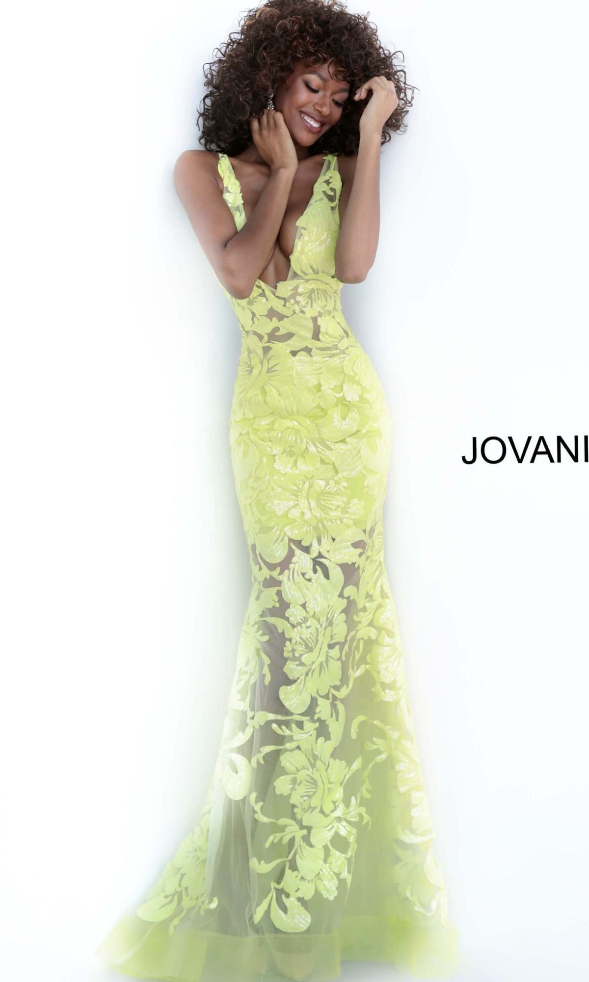 A woman in a sleeveless, sheer yellow Jovani Plus-Size Sheer Prom Dress with Sequins smiles against a white background, with the