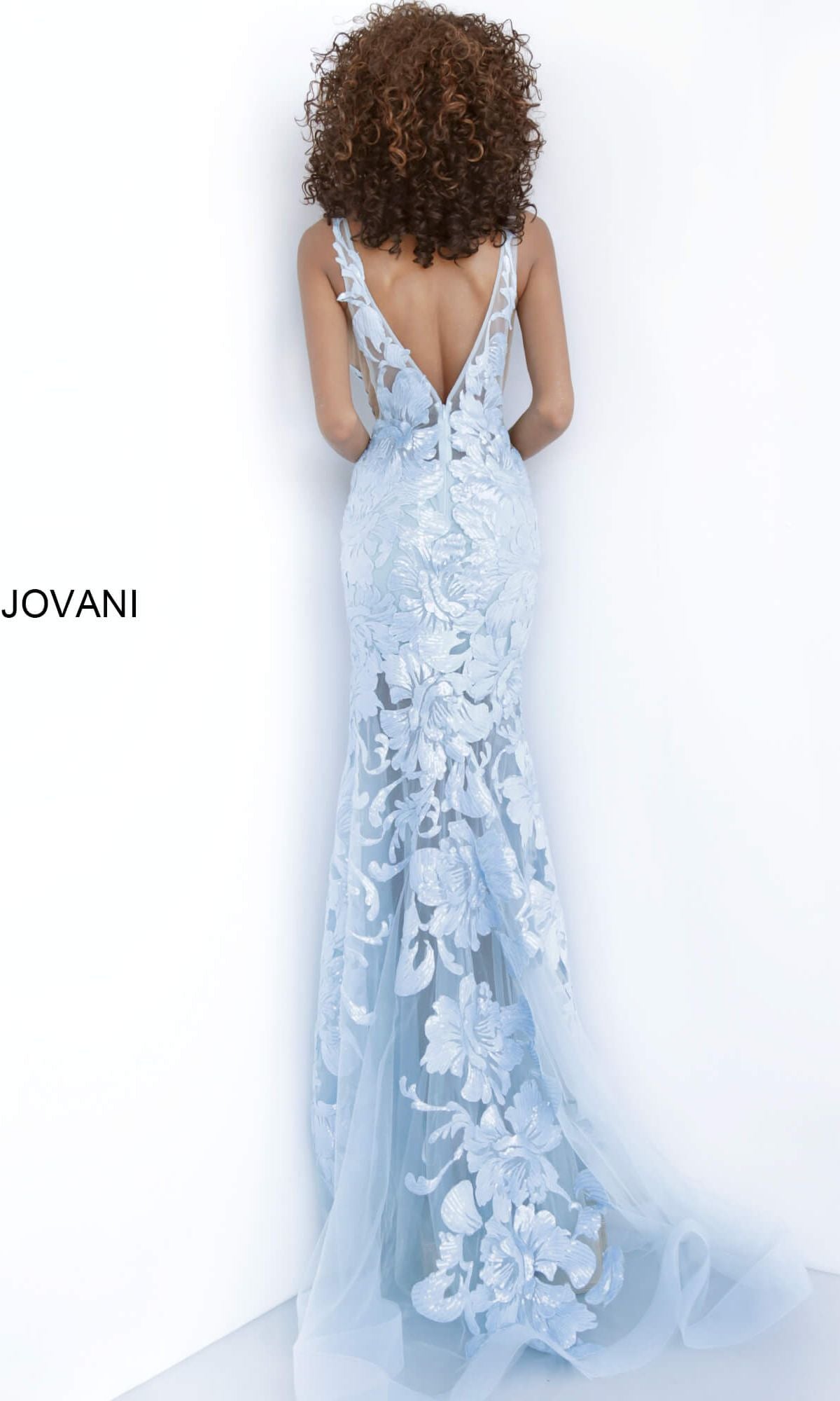 A woman with curly hair faces a white wall, wearing a light blue, floor-length Jovani Embroidered Long Prom Dress with sequins and a backless design.
