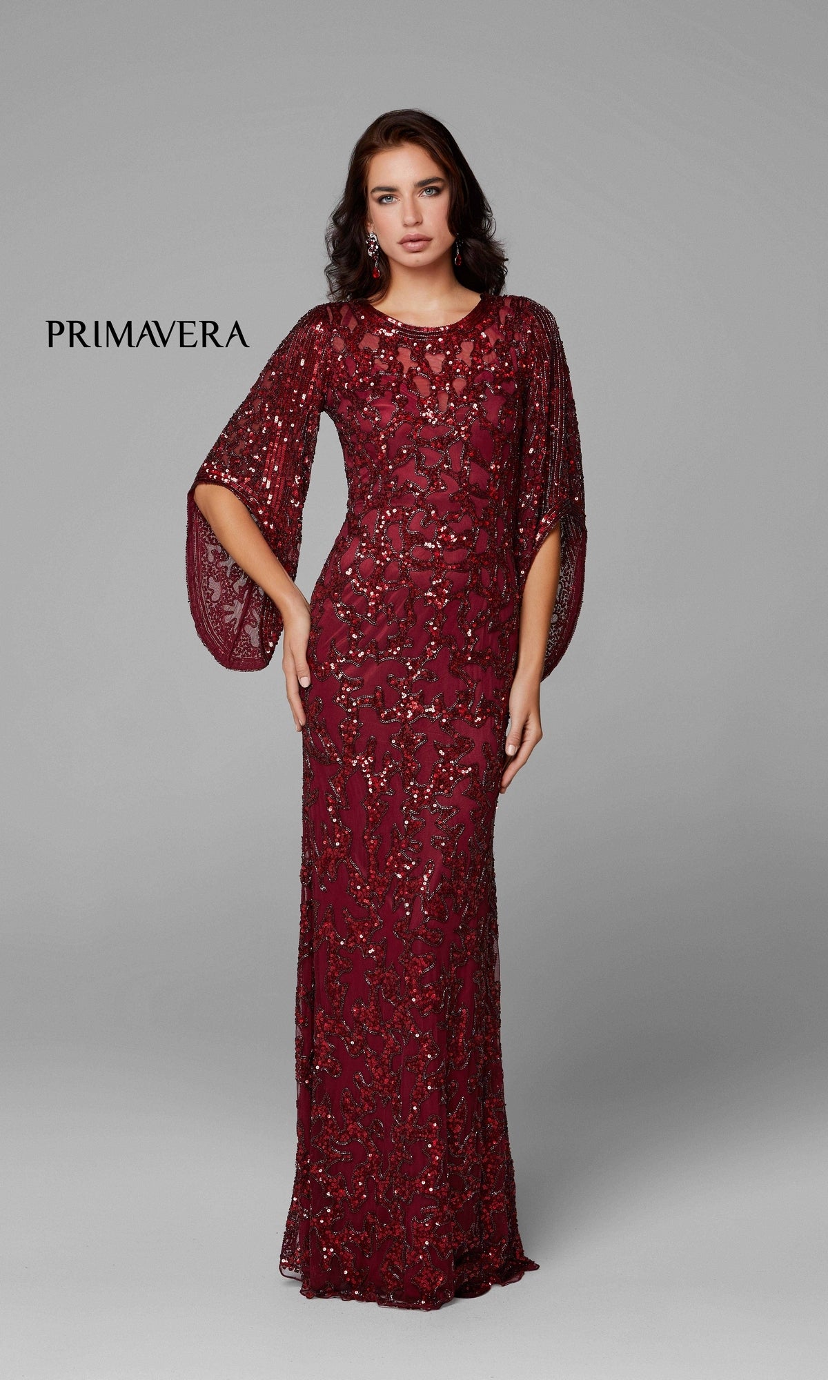 A woman poses in the Long Bell Sleeve Beaded Evening Gown 9713—floor-length, burgundy, and adorned with beads—featuring flowing bell sleeves against a plain gray background.