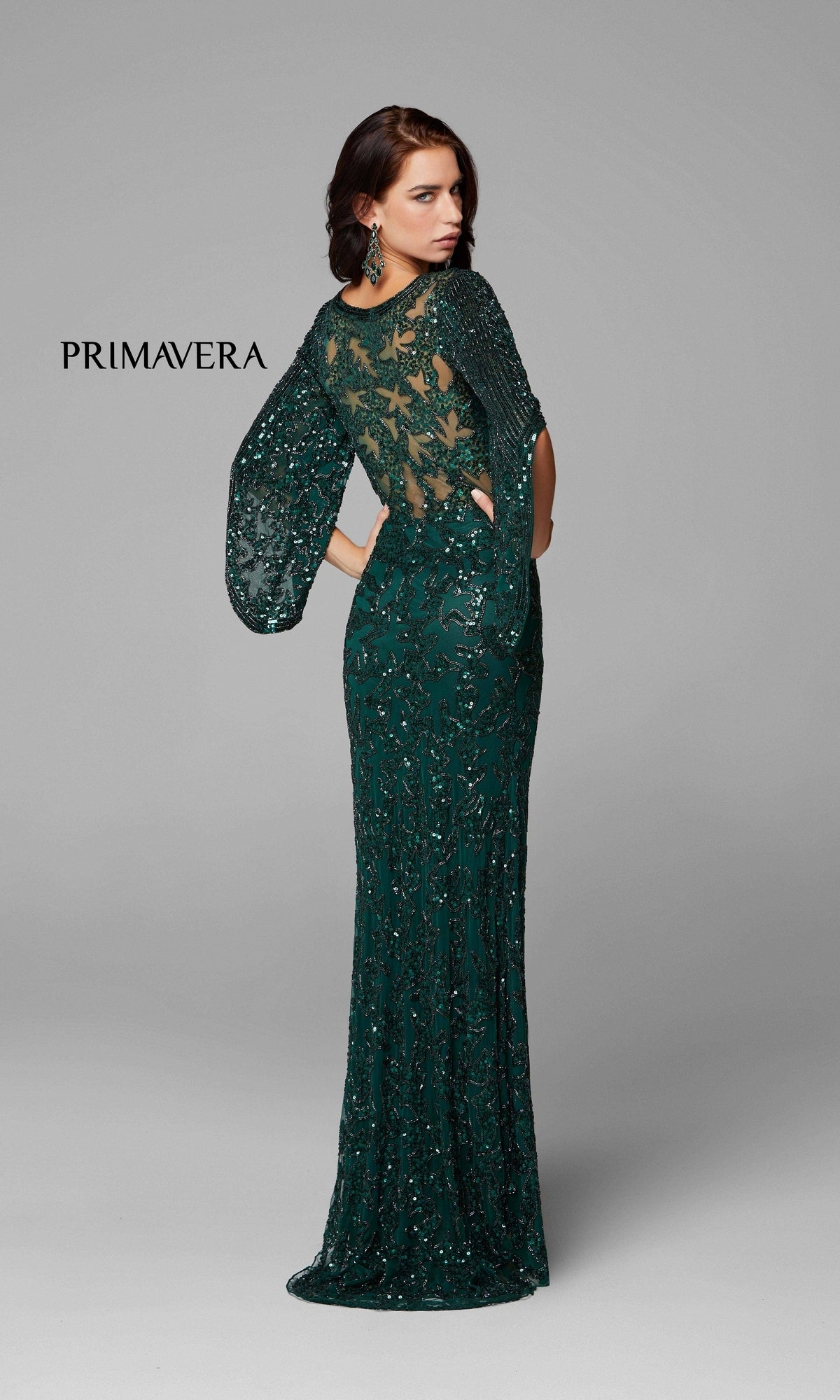 A woman models the Long Bell Sleeve Beaded Evening Gown 9713—a floor-length, dark green dress with sheer bell sleeves and a partially transparent back. The plain light gray background features