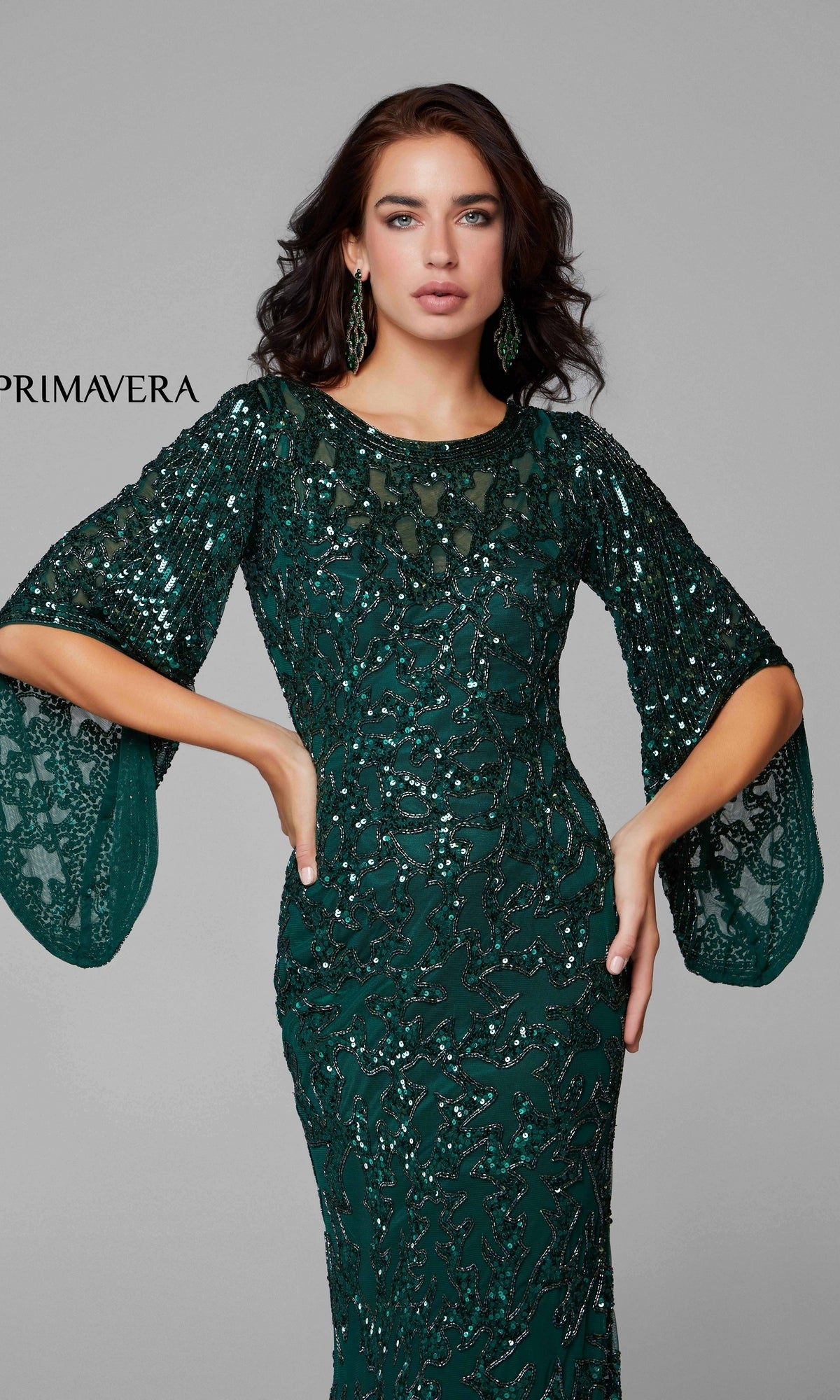 A woman poses with one hand on her hip, wearing the Long Bell Sleeve Beaded Evening Gown 9713 in emerald green against a plain gray background.