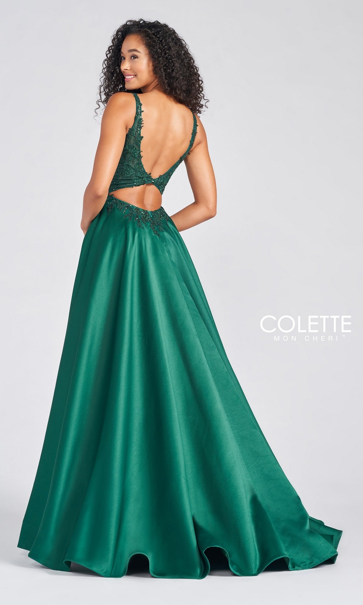 A woman in a floor-length, sleeveless emerald green V-Neck Satin Prom Dress by Colette by Daphne with a low back and lace details stands facing slightly away from the camera.
