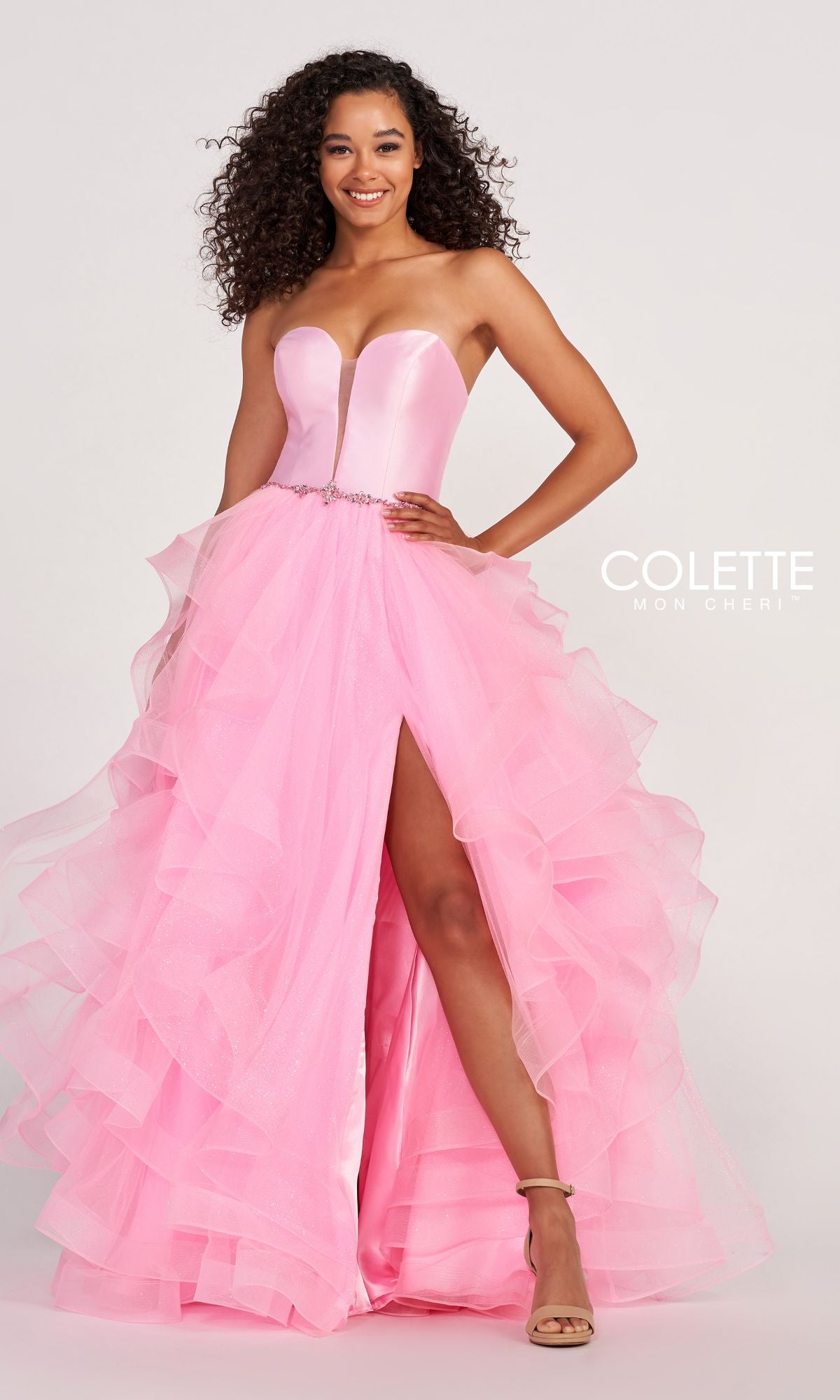 A woman wearing the Colette Glitter-Tulle Layered Long Prom Ball Gown in bright pink, featuring ruffled layers and a thigh-high slit, stands smiling against a plain background.