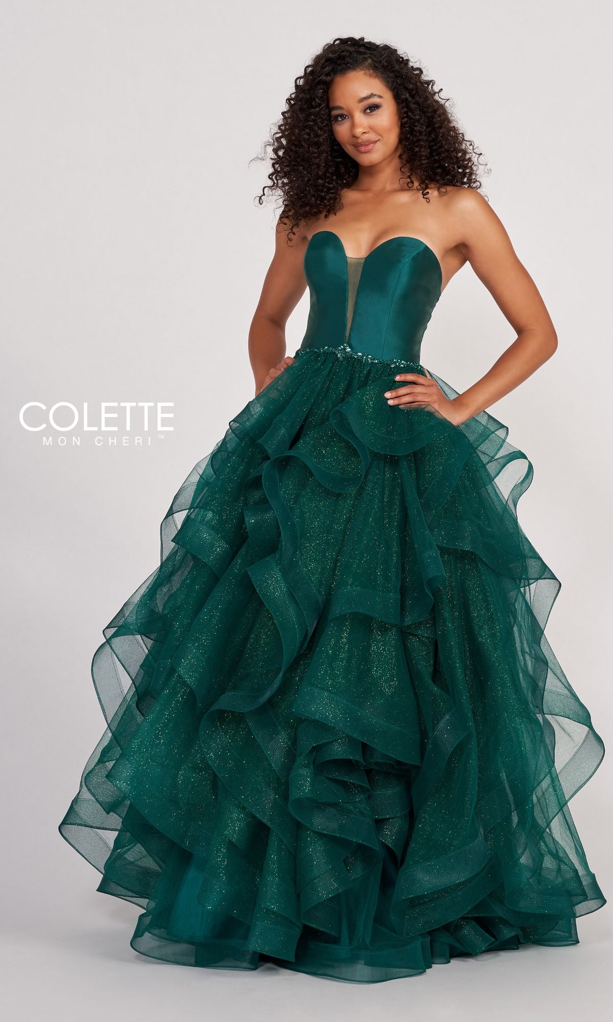 A woman poses in the Colette Glitter-Tulle Layered Long Prom Ball Gown—an emerald green, strapless ball gown with a fitted bodice, ruffled skirt, subtle shimmer, and a sheer front panel.
