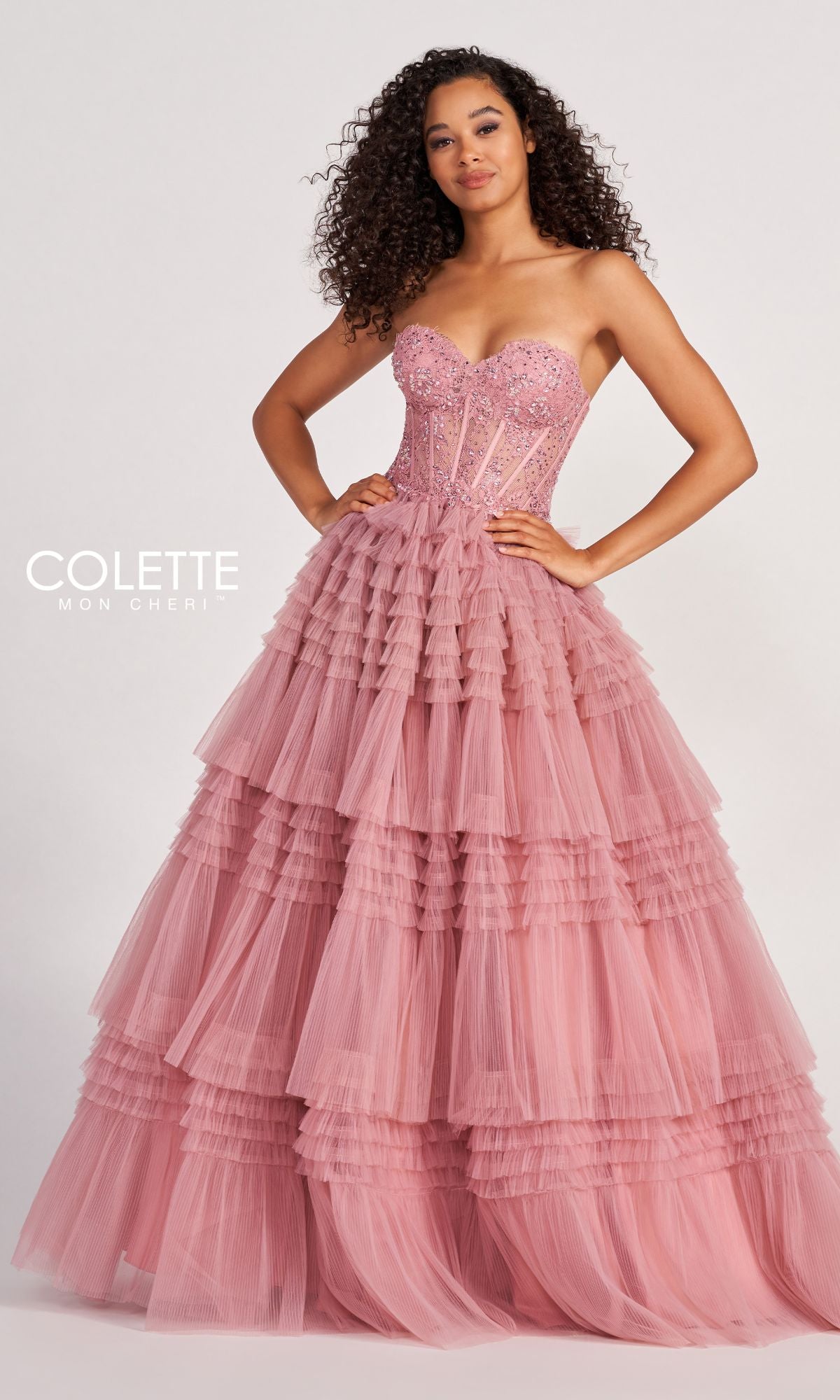 A woman wears the Colette CL2017 Strapless Ruffled Prom Ball Gown, featuring a beaded corset bodice and layered pink tulle skirt, standing elegantly against a plain background.
