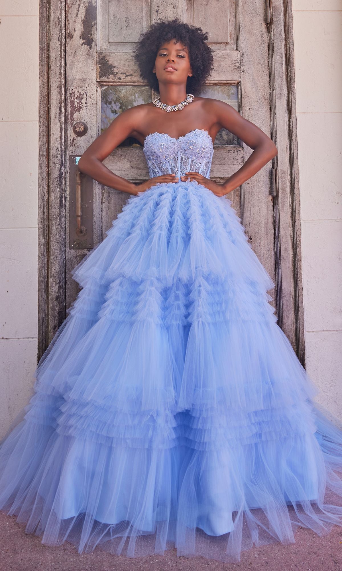 A woman poses before a weathered wooden door wearing the Colette CL2017 Strapless Ruffled Prom Ball Gown, which features a beaded corset bodice, layered light blue tulle skirt, and a statement necklace.