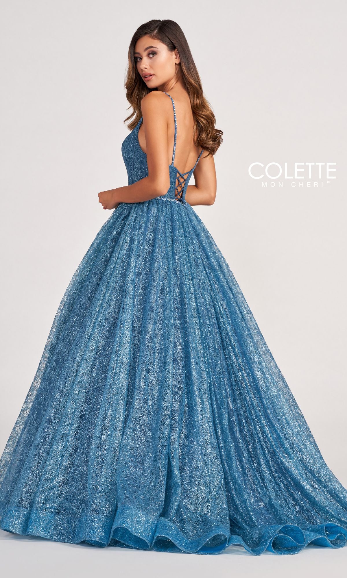 A woman wears the Colette CL2018 Deep V-Neck Glitter-Lace Ball Gown, featuring a fitted bodice, lace-up back, and full skirt, standing against a plain light background. The