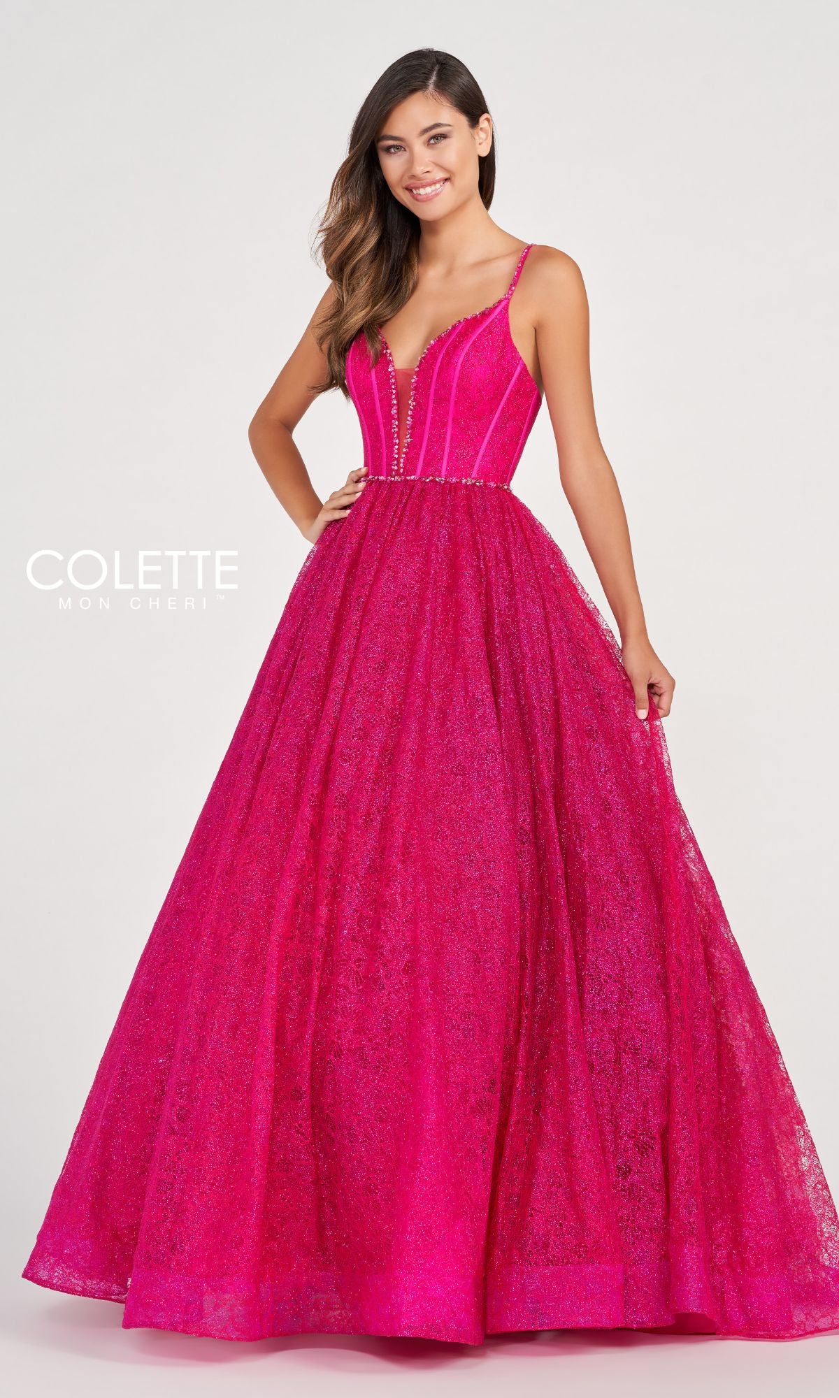 A woman poses against a plain light background in the Colette CL2018 Deep V-Neck Glitter-Lace Ball Gown—bright pink, floor-length, with a fitted bodice, spaghetti straps, and textured lace overlay.