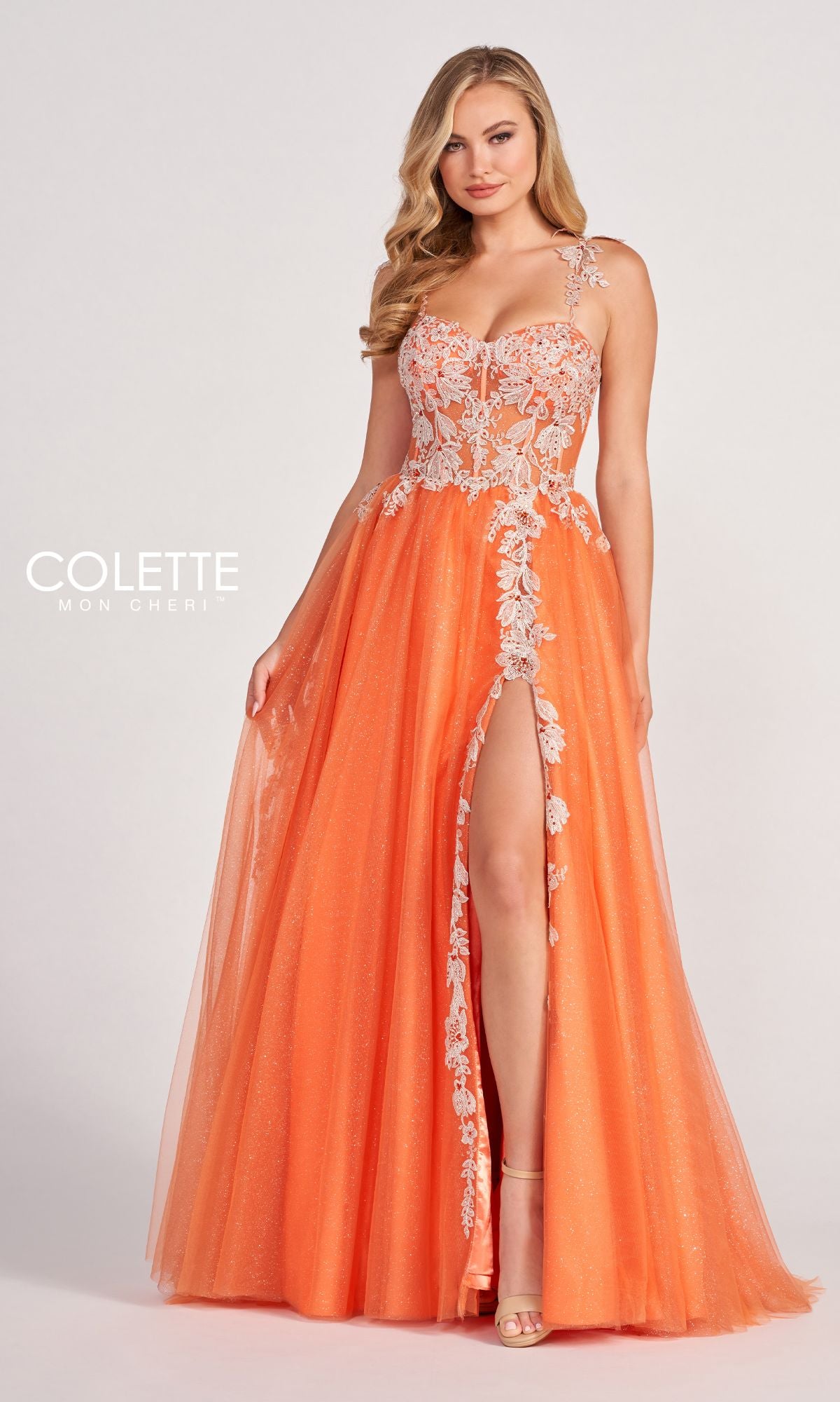 A woman models the Colette CL2020 Glitter-Lace Long A-Line Prom Dress in vibrant orange, showcasing a floor-length skirt with a high slit, floral lace bodice, and thin straps against a plain light background.