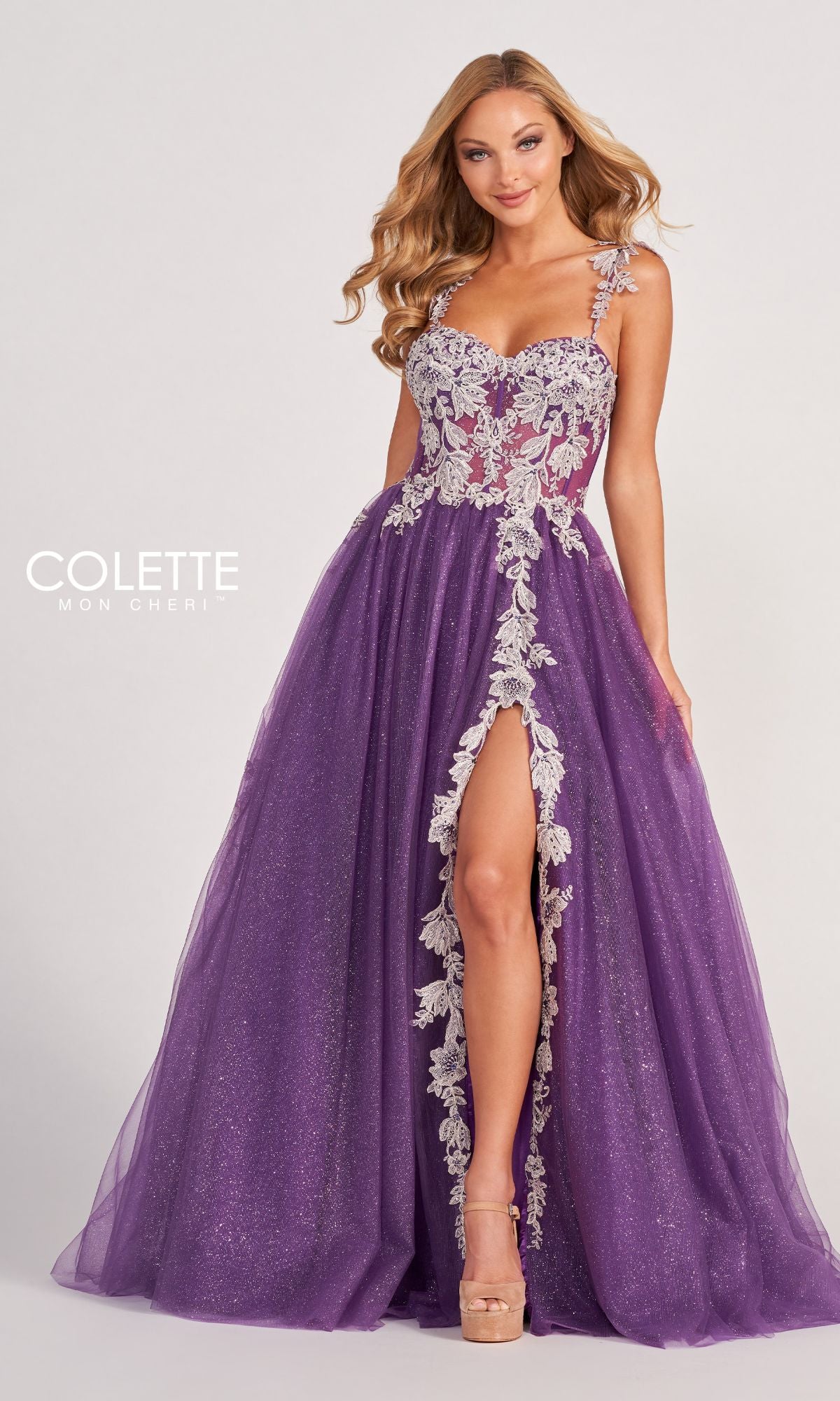 A woman wears the Colette CL2020 Glitter-Lace Long A-Line Prom Dress, featuring a sleeveless purple design with a lace bodice, floral appliqué straps, high slit, and glamorous glittery tulle skirt.
