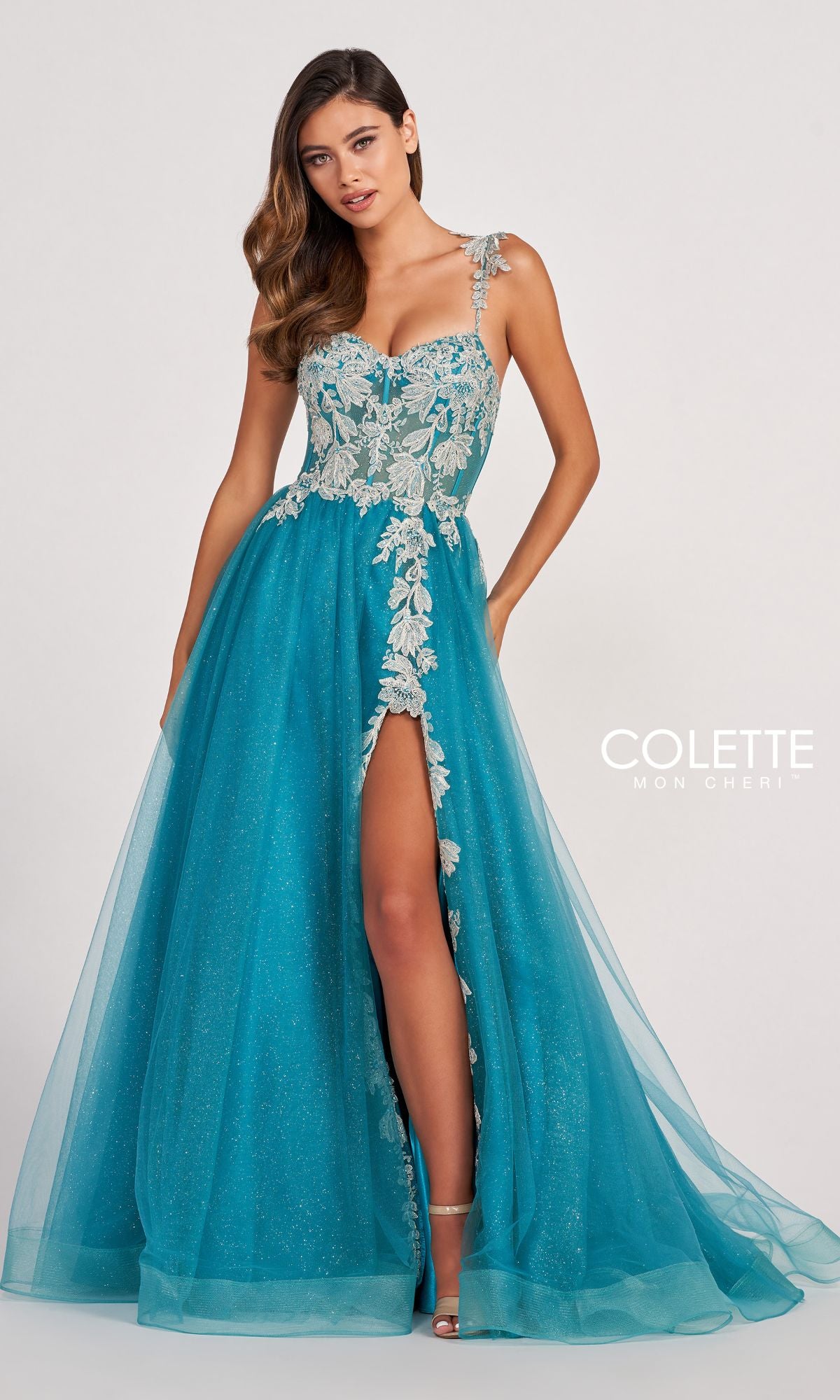 A woman wears the Colette CL2020 Glitter-Lace Long A-Line Prom Dress, a teal designer gown with silver floral embroidery, sweetheart neckline, one-shoulder strap, and high side slit—perfect for special occasions.