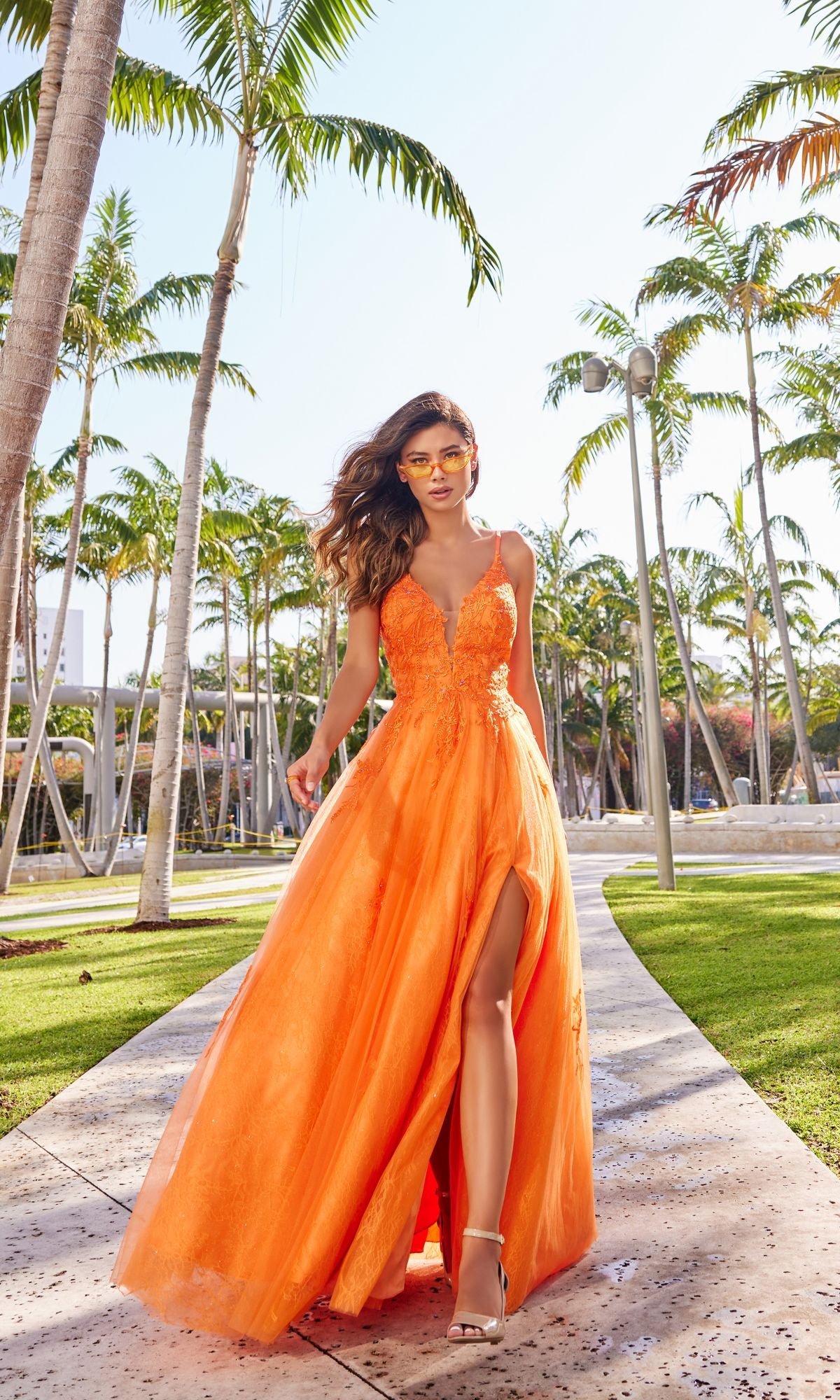 A woman wearing the Beaded Lace Long A-Line Prom Dress Colette CL2025 walks outdoors along a palm tree-lined path on a sunny day.