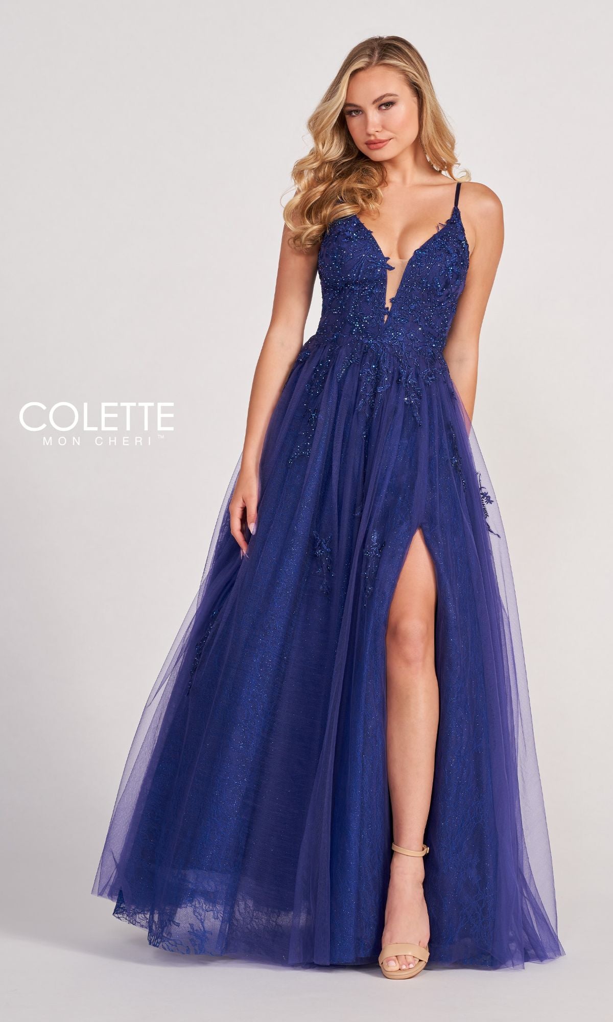 A woman wearing the Beaded Lace Long A-Line Prom Dress Colette CL2025, featuring a deep blue hue, floor-length slit, thin straps, floral embellishments, beaded lace accents, and tulle overlay stands against a plain background.