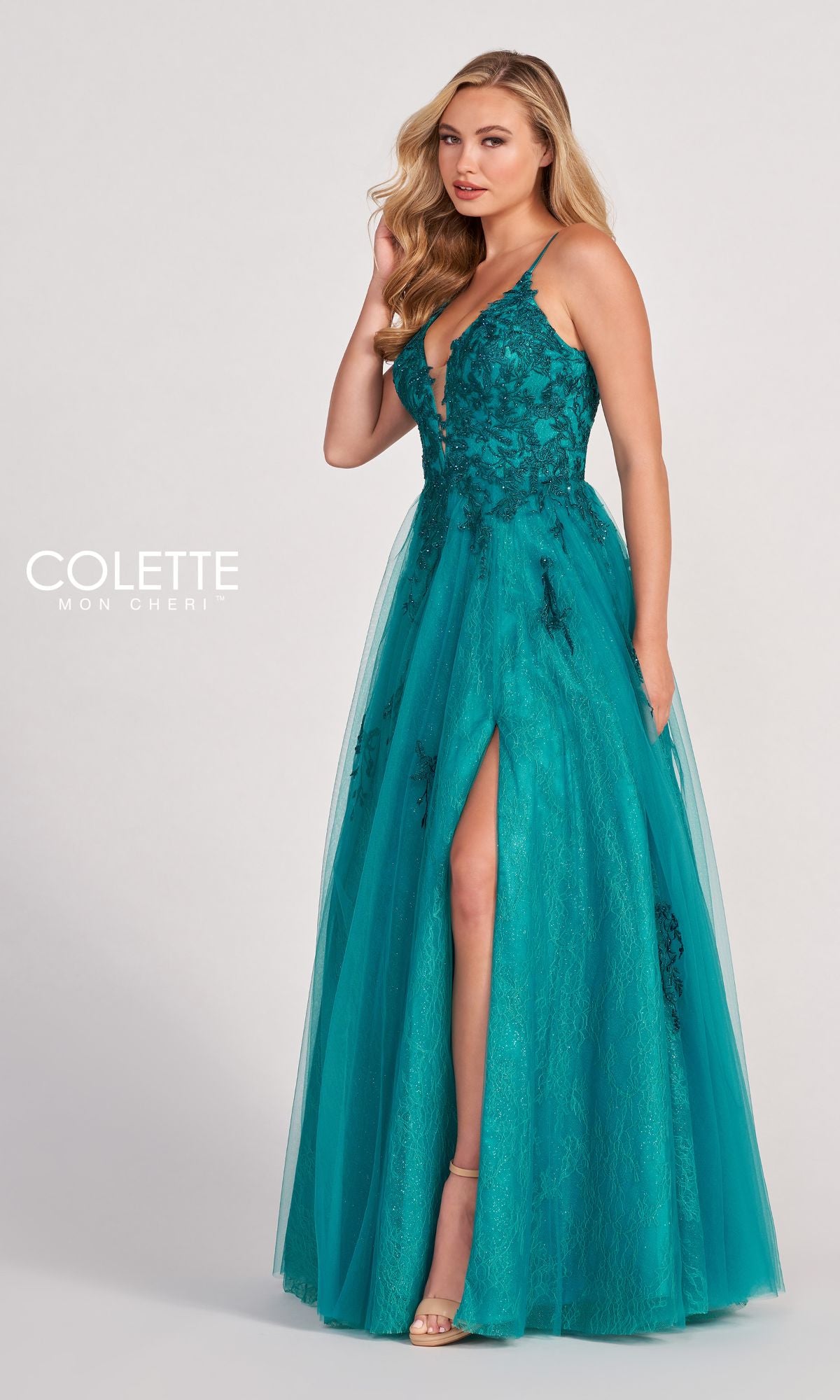 A woman wears the Beaded Lace Long A-Line Prom Dress Colette CL2025, featuring teal fabric, thin straps, a deep V-neckline, beaded lace detailing, and a thigh-high slit, as she stands against a plain background.