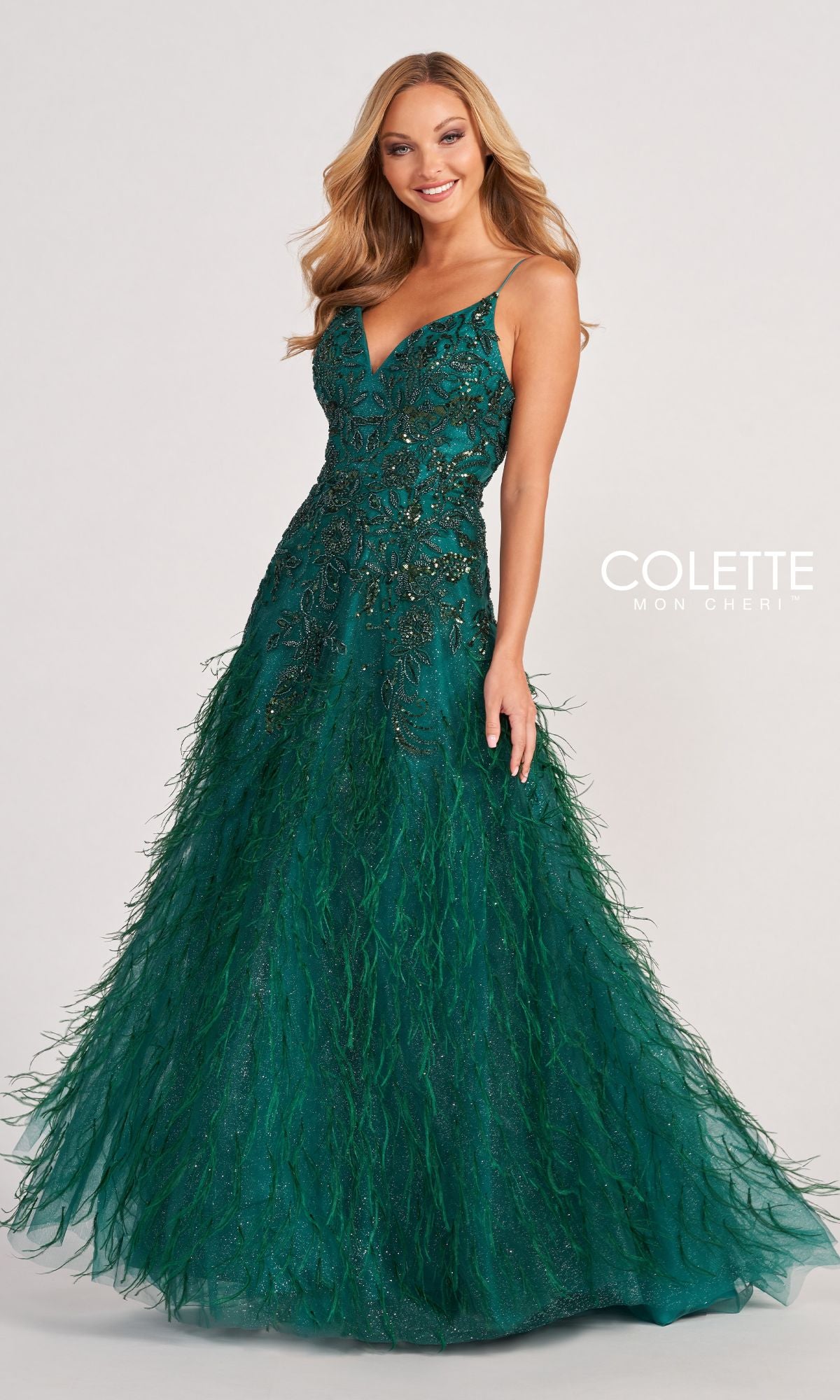 A woman stands against a plain background in the Colette CL2044 Feather Prom Ball Gown, a deep green beaded dress with a fitted bodice and flowing feathered floor-length skirt, radiating elegant evening glamour.