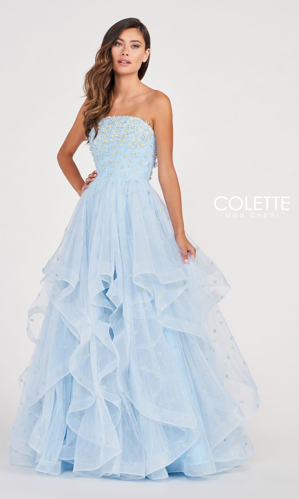 A woman stands against a plain white background wearing the Colette CL2055 Strapless Daisy Ball Gown—a light blue, ruffled-layered dress with bead embellishments.