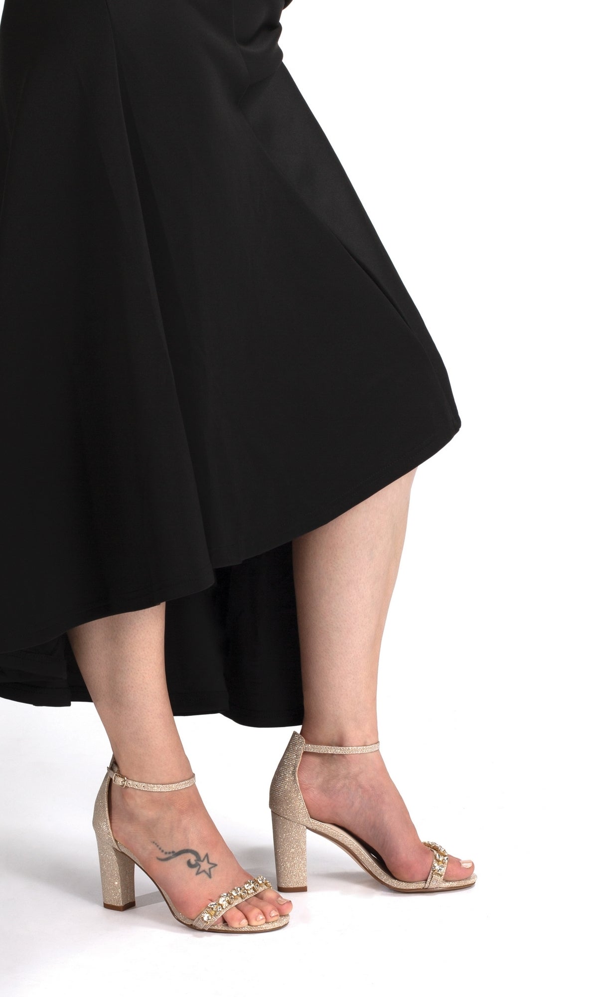 Wearing a black dress and Camila Champagne 2.75in Prom Shoes by Touch Ups 4522 with rhinestone heels, a person displays a star and swirl ankle tattoo while standing against a white background.