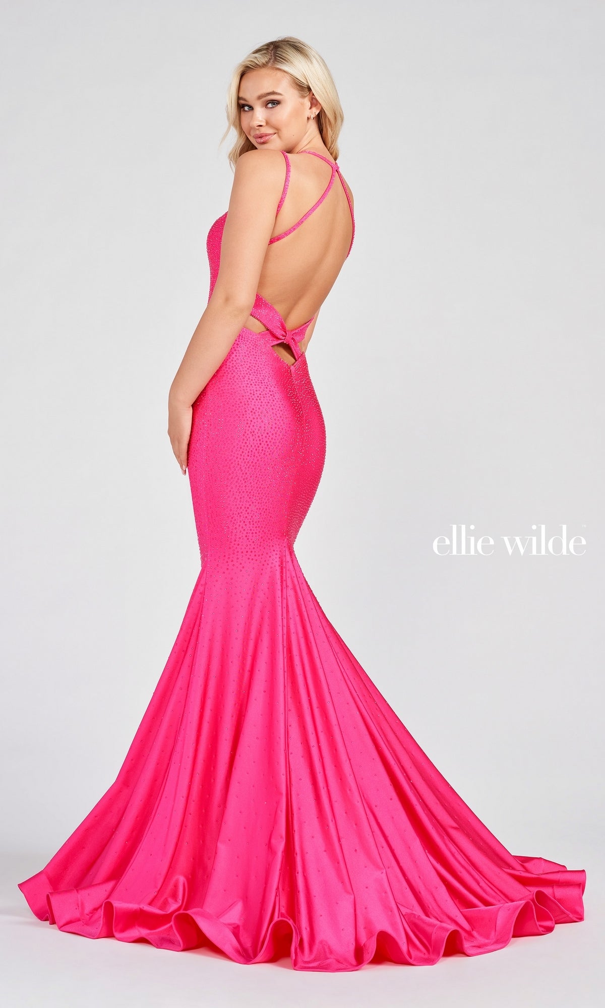 A woman poses in a bright pink Strappy-Back Long Beaded Ellie Wilde Formal Gown—a backless, floor-length mermaid dress with thin straps and a small bow—against a plain background. The brand