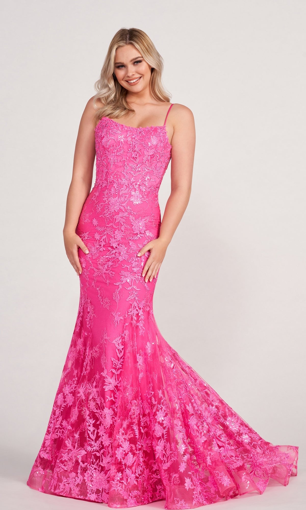A woman smiles, standing in front of a plain background, wearing the Ellie Wilde Embroidered-Lace Mermaid Prom Dress in bright pink with thin straps and elegant lace detailing.