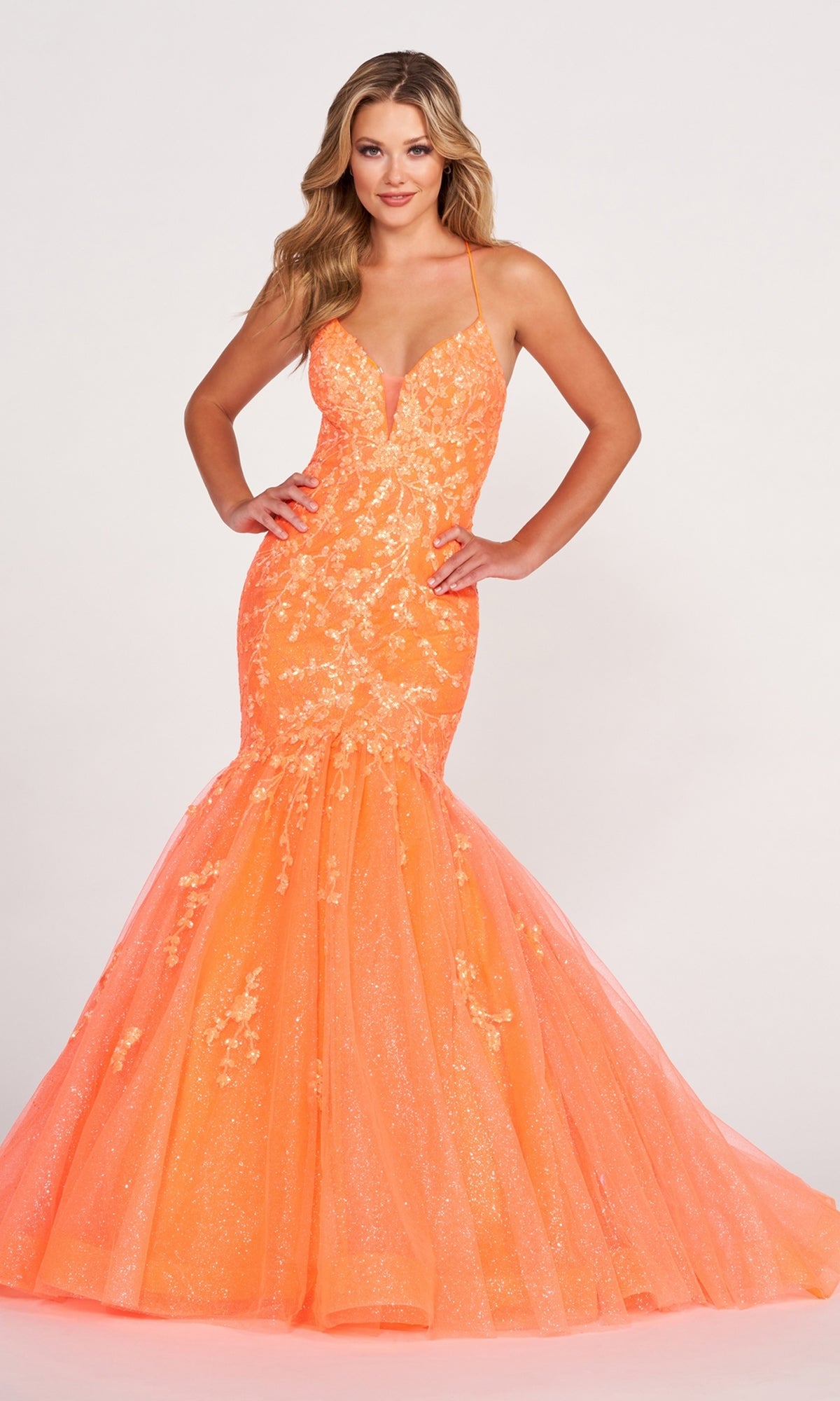 A woman poses against a plain white background in the Ellie Wilde Glitter-Tulle Long Mermaid Prom Gown, featuring a bright orange hue, sleeveless design, and floral lace with sequin details.
