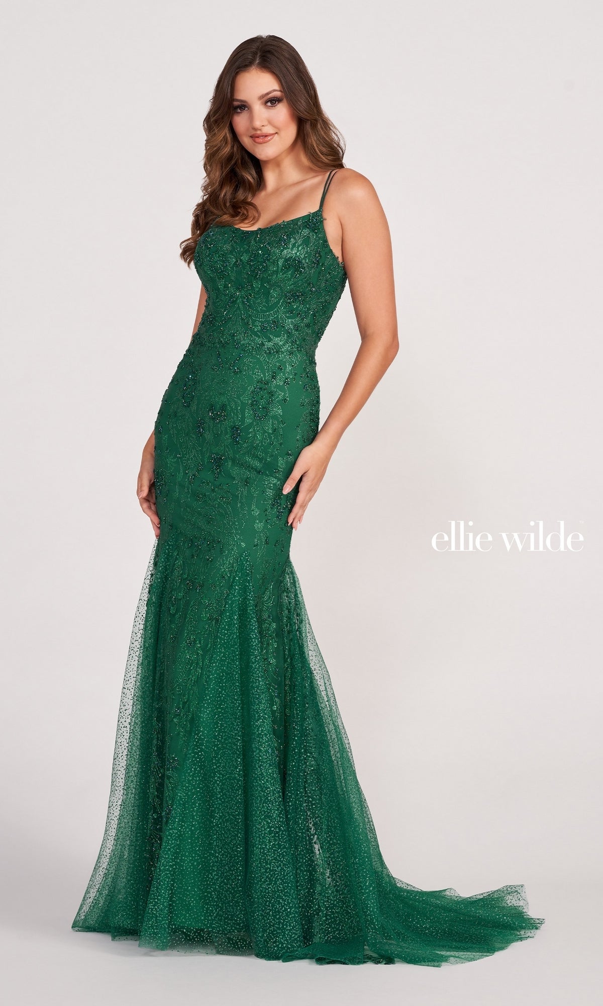 A woman models the Glitter-Jersey Long Mermaid Ellie Wilde Prom Gown—an elegant, fitted floor-length dress in green with thin straps and sequin details. She stands against a plain background with