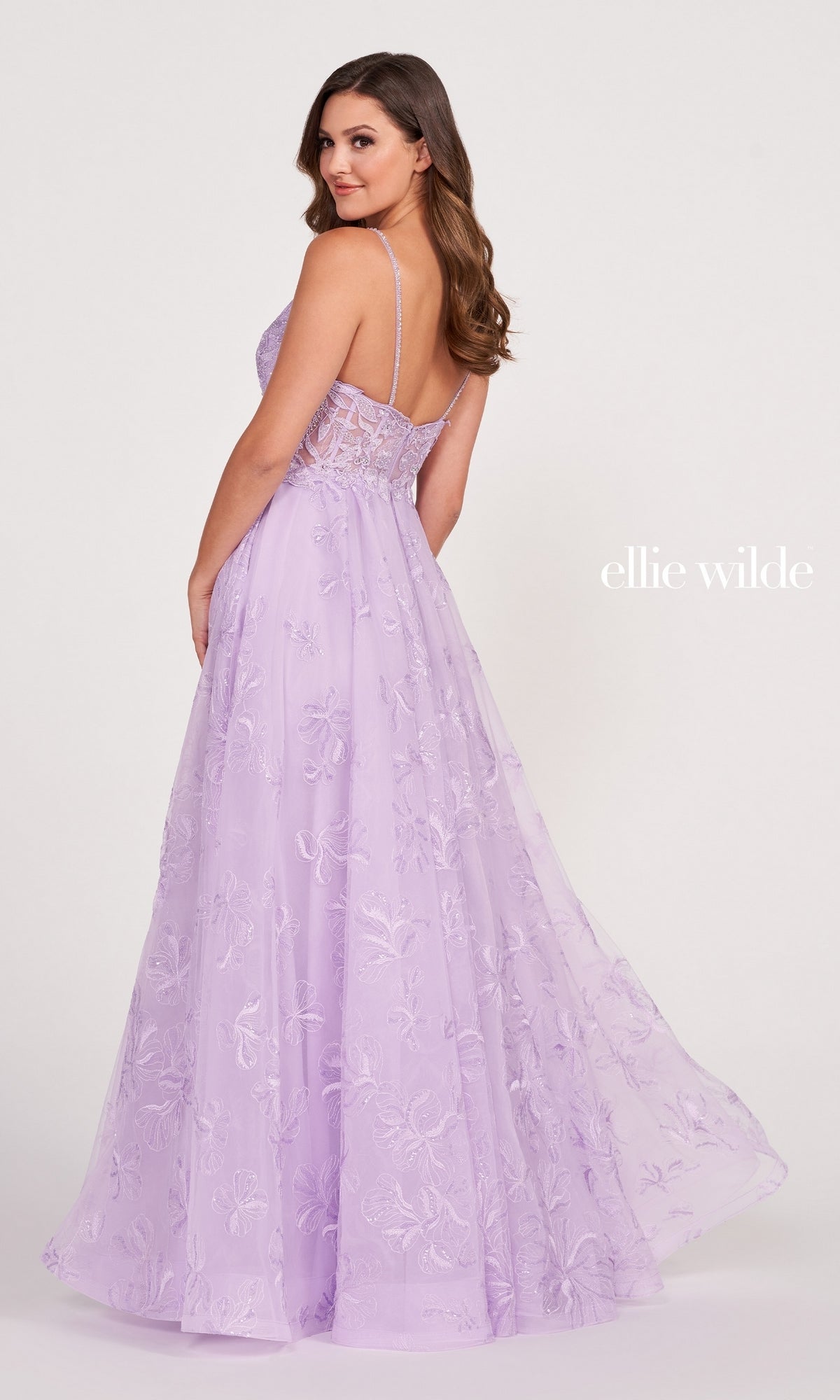 A woman faces backward, looking over her shoulder in the Tulle Embroidered A-Line Prom Dress by Ellie Wilde—a lavender, floor-length gown with thin straps, a sheer embellished bodice, and floral embroidery.