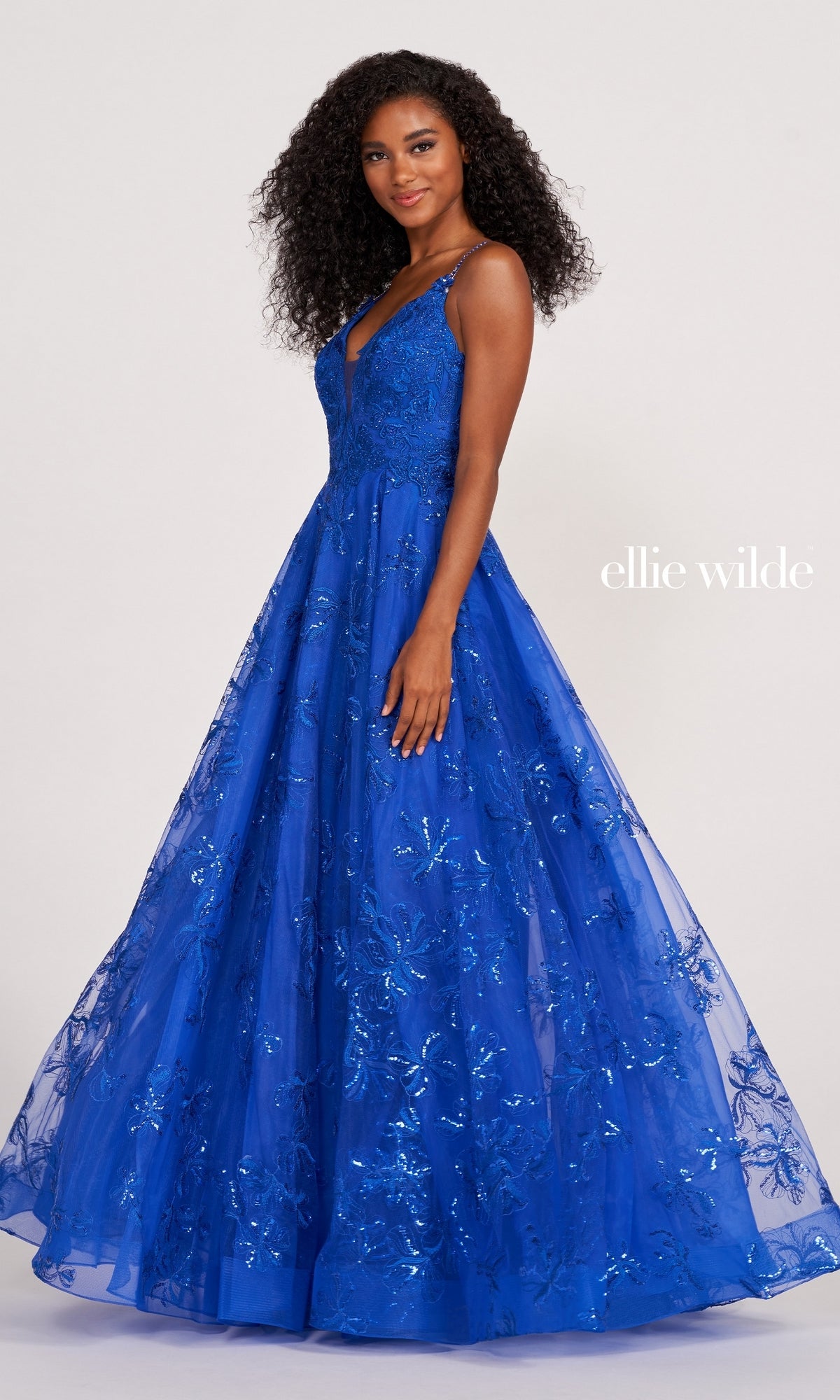 Woman in a floor-length royal blue Tulle Embroidered A-Line Prom Dress by Ellie Wilde, featuring glittery floral patterns and sleeveless design, stands against a plain background. The text