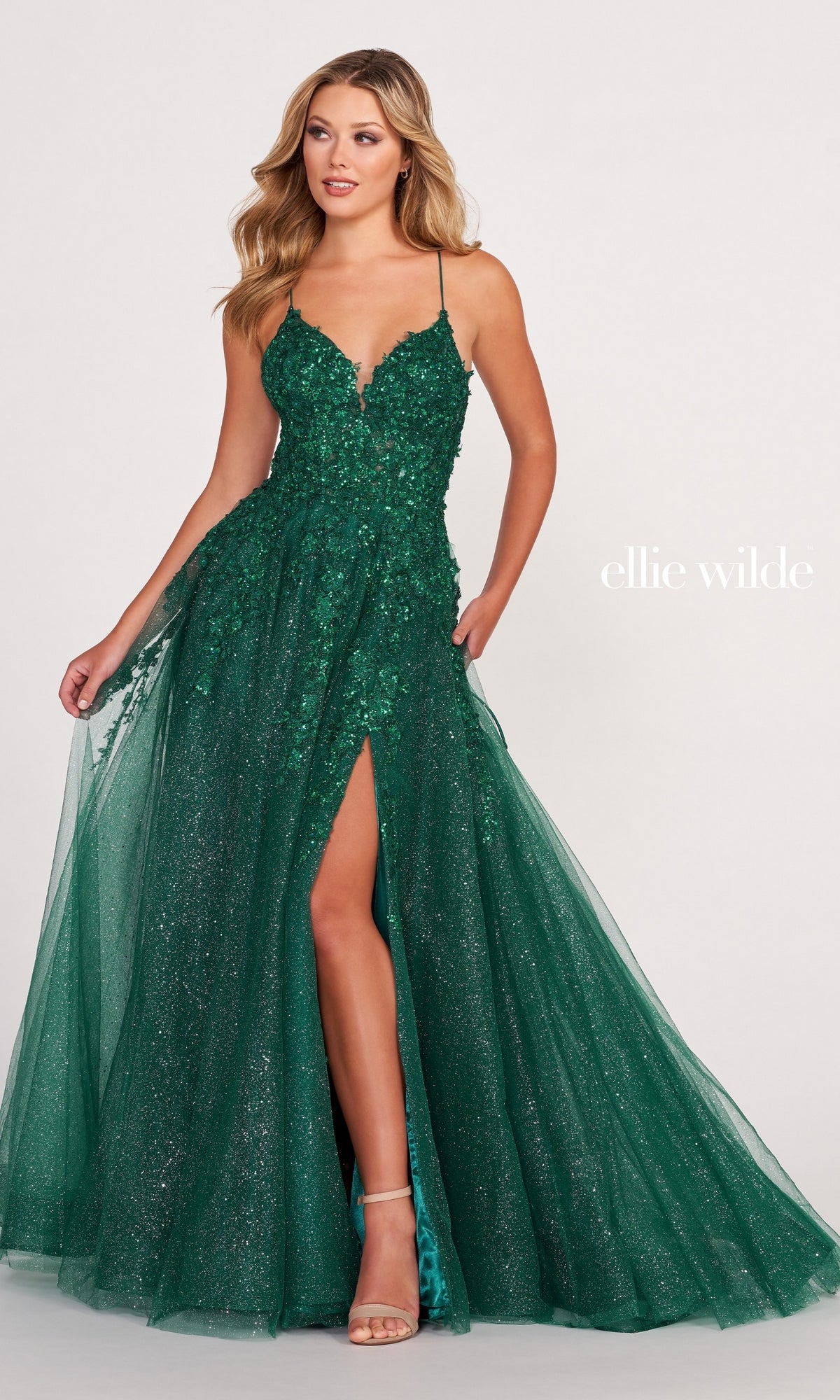 A woman models the Ellie Wilde Backless Designer Prom Dress EW34053, a sparkling emerald green ball gown with a deep neckline, thin straps, floral details, thigh-high slit, and tulle overlay. The plain background highlights the dazzling dress.