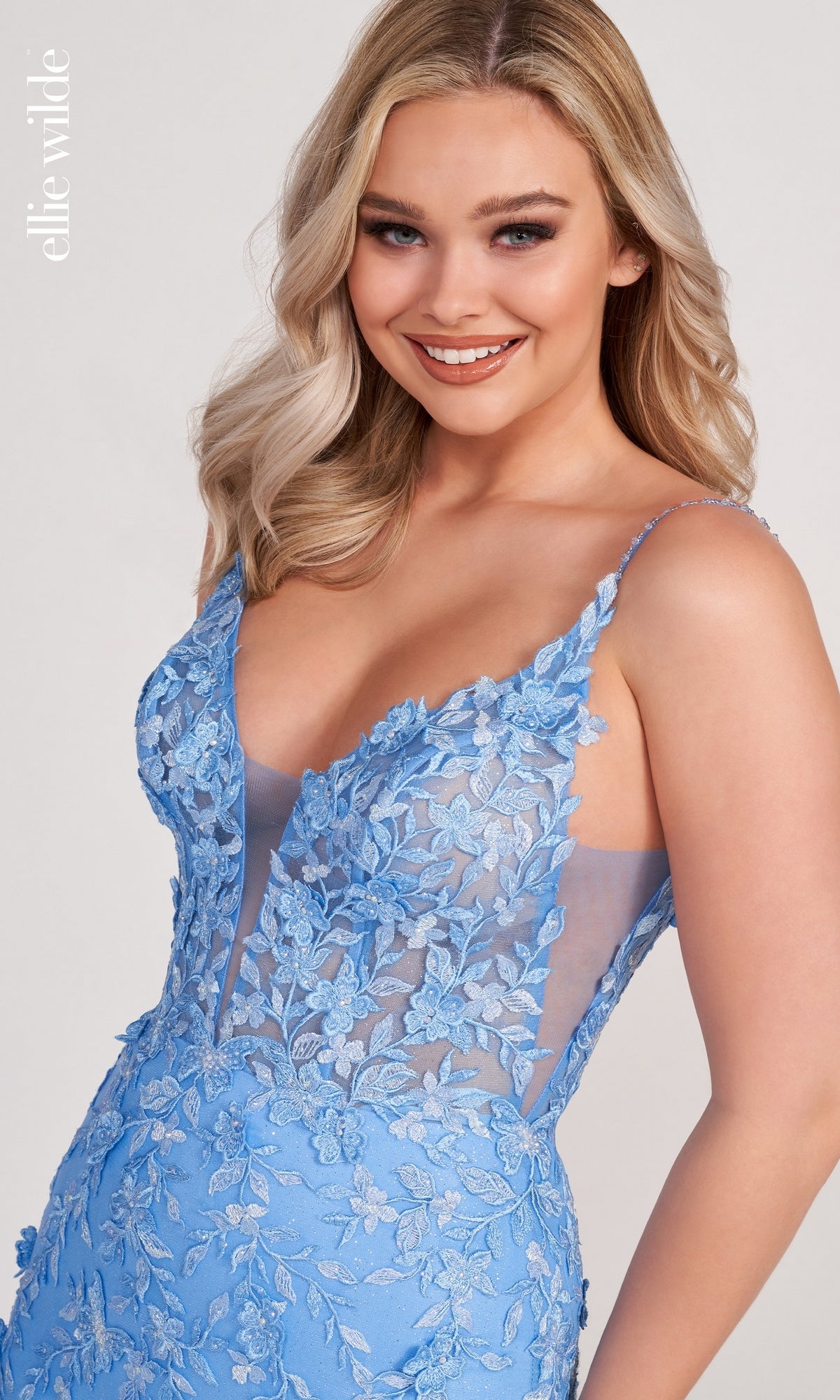 A woman with long blonde hair smiles at the camera, wearing a Lace Embroidered Mermaid Gown With Sheer Bodice in light blue, adorned with floral patterns, sheer panels, and thin straps.