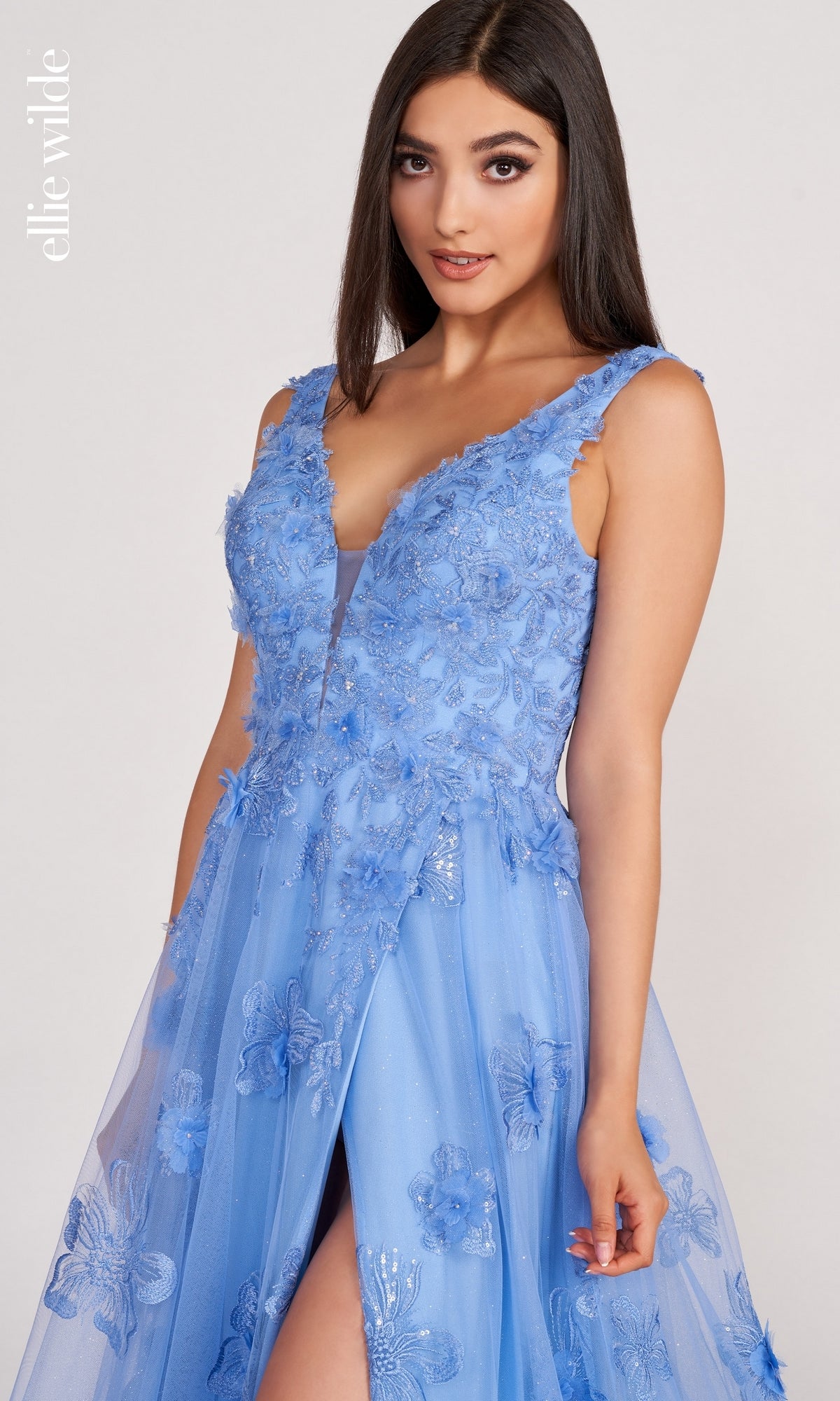 A woman wears the Ellie Wilde Faux-Wrap Embroidered-Lace Prom Dress, a sleeveless light blue gown with embroidered lace, a sheer overlay, and a front slit, standing against a plain background.