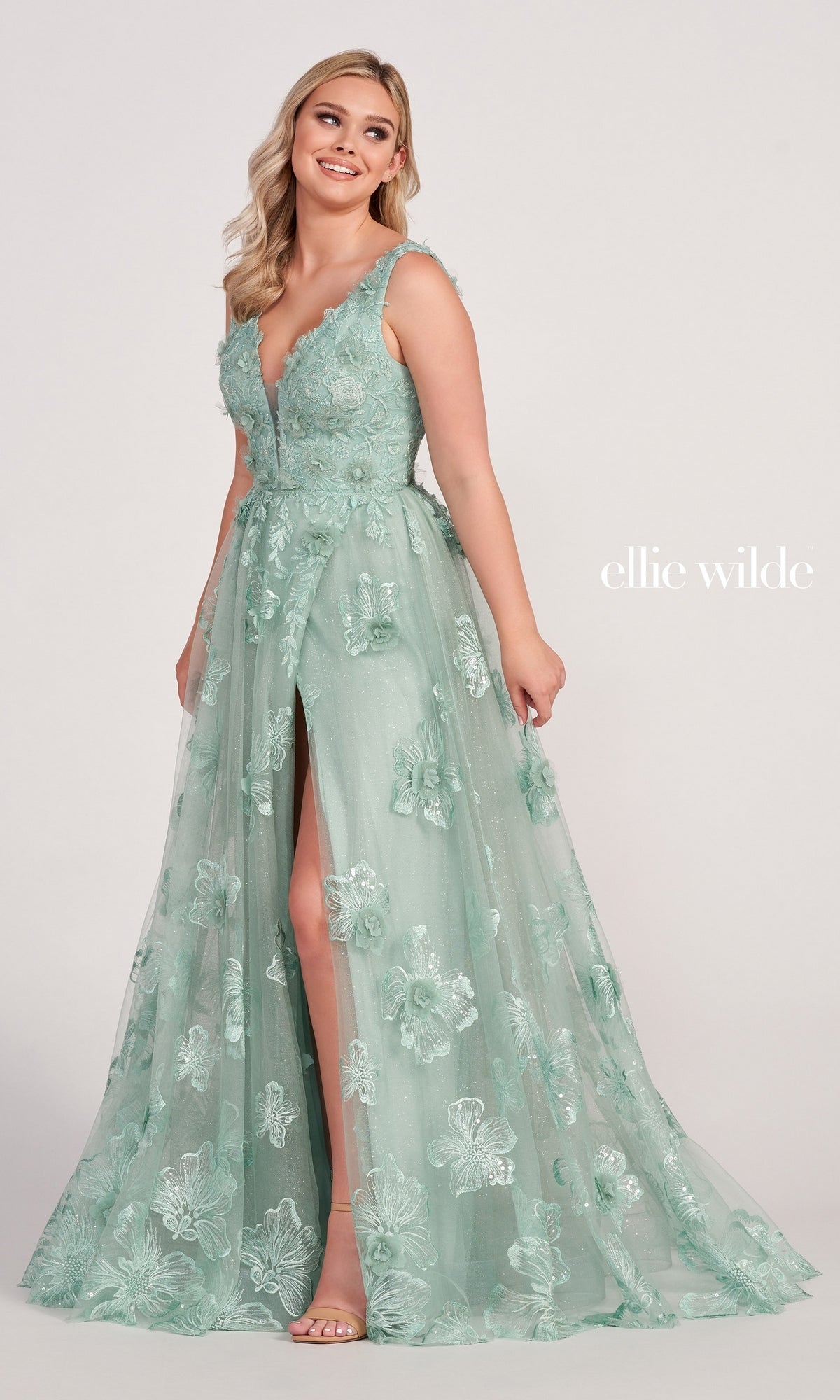 A woman in the Ellie Wilde Faux-Wrap Embroidered-Lace Prom Dress, featuring a sleeveless mint green design with a deep V-neckline and high slit, stands against a plain background.