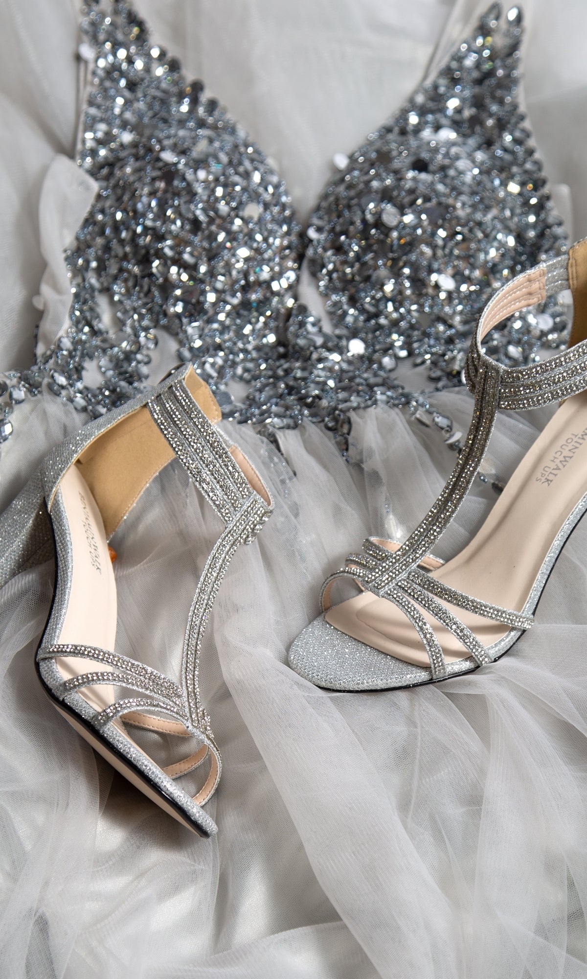 The Silver Block-Heel Prom Shoes by Touch Ups 4526, featuring rhinestones and elegant straps, rest on a silver sequined tulle dress for a stunning prom night look.